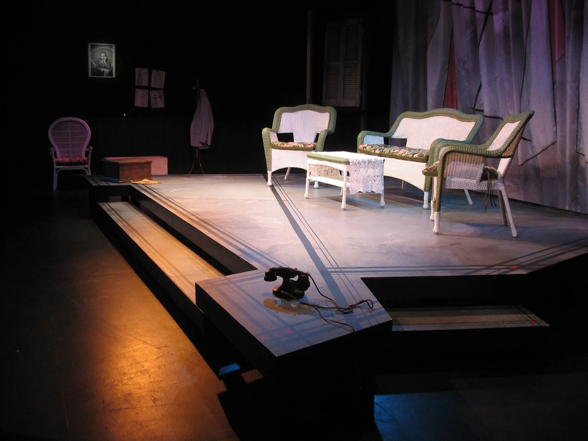 The image shows a stage set with vintage furniture, including a green and white sofa set, a pink chair, and a small table with lace cloth. There is a black rotary phone on a block in the foreground, and dark curtains with a purple curtain in the back
