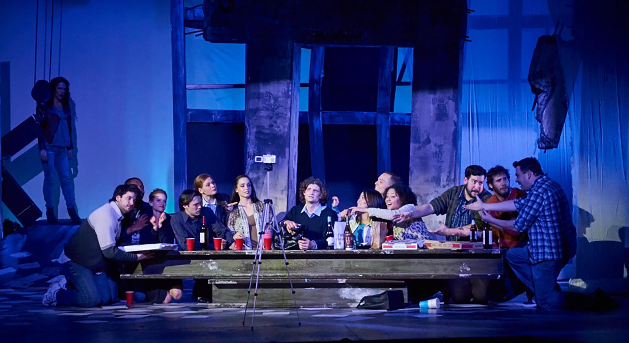 A theatrical stage scene depicting multiple actors sitting and kneeling around a table, with some reaching out their hands. The set includes a wooden structure and hanging fabric, with a woman standing on the left side.