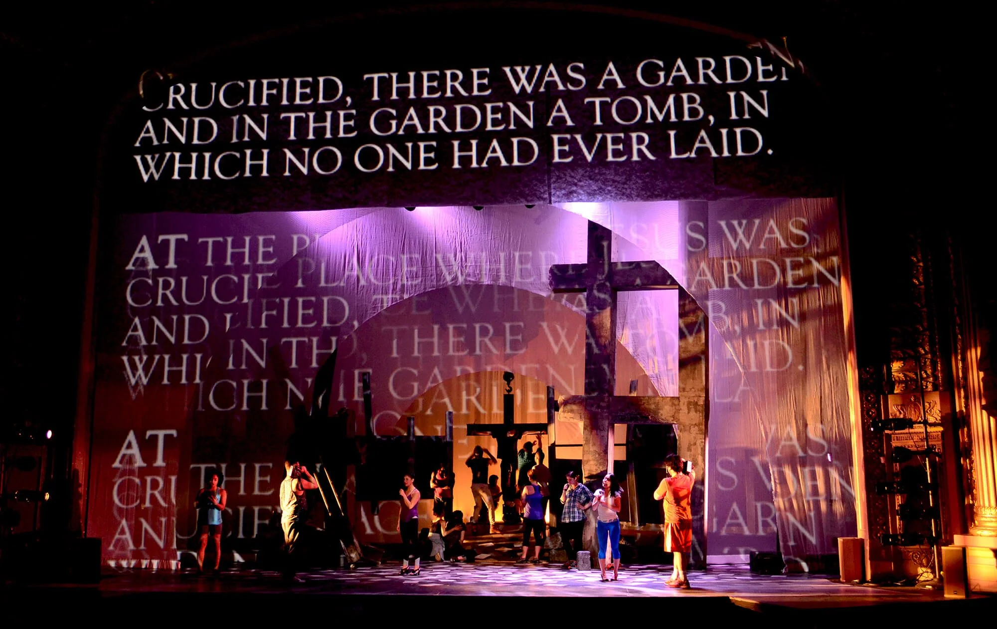Stage with people rehearsing, large projected text about the crucifixion, with silhouettes of crosses and ancient ruins and purple, orange, and yellow lighting.