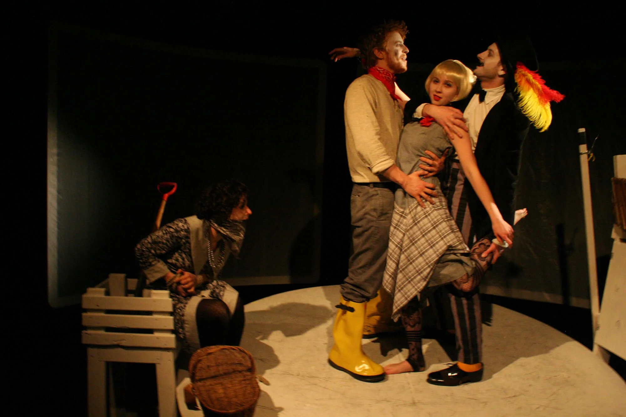 Actors performing a scene on stage with three characters in the center, one person holding another in a hug, and a person sitting on a bench with a shovel behind them, in a theatrical production.