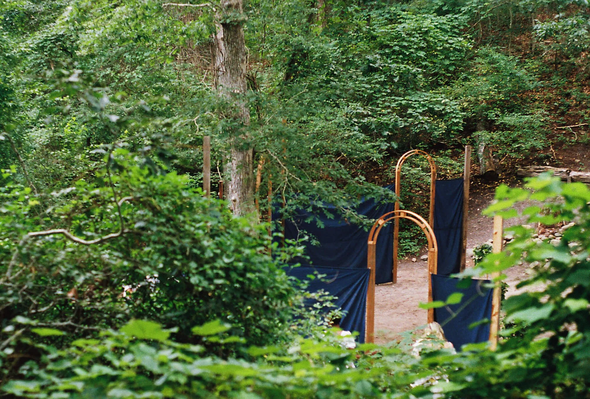 A pathway through a green wooded area with blue fabric panels and wooden frames, possibly part of a temporary outdoor installation or event, with trees and leaves surrounding it.