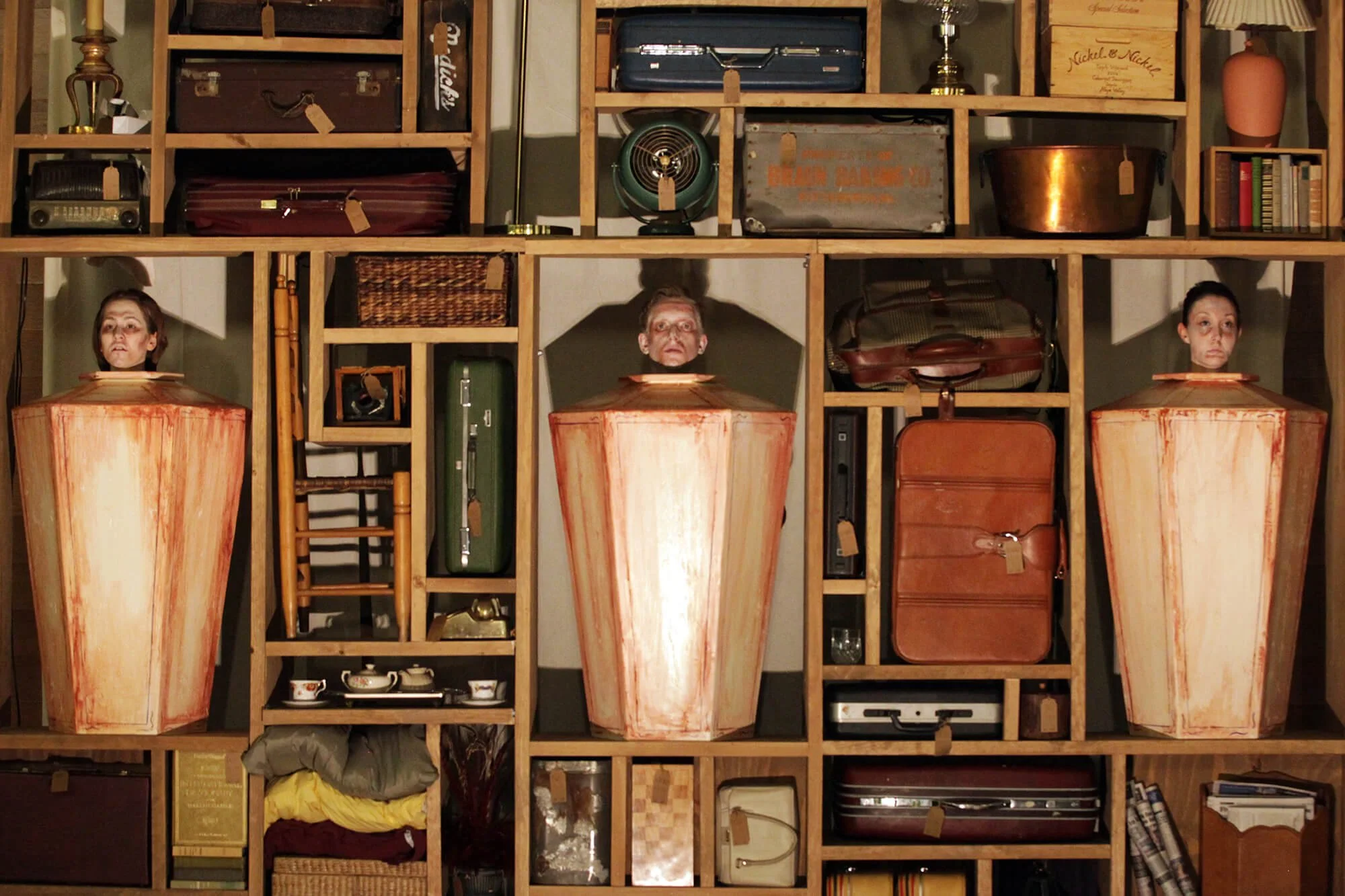 Three people with their heads in large, open-mouthed wooden boxes, behind a grid of wooden shelves filled with various vintage and decorative items.