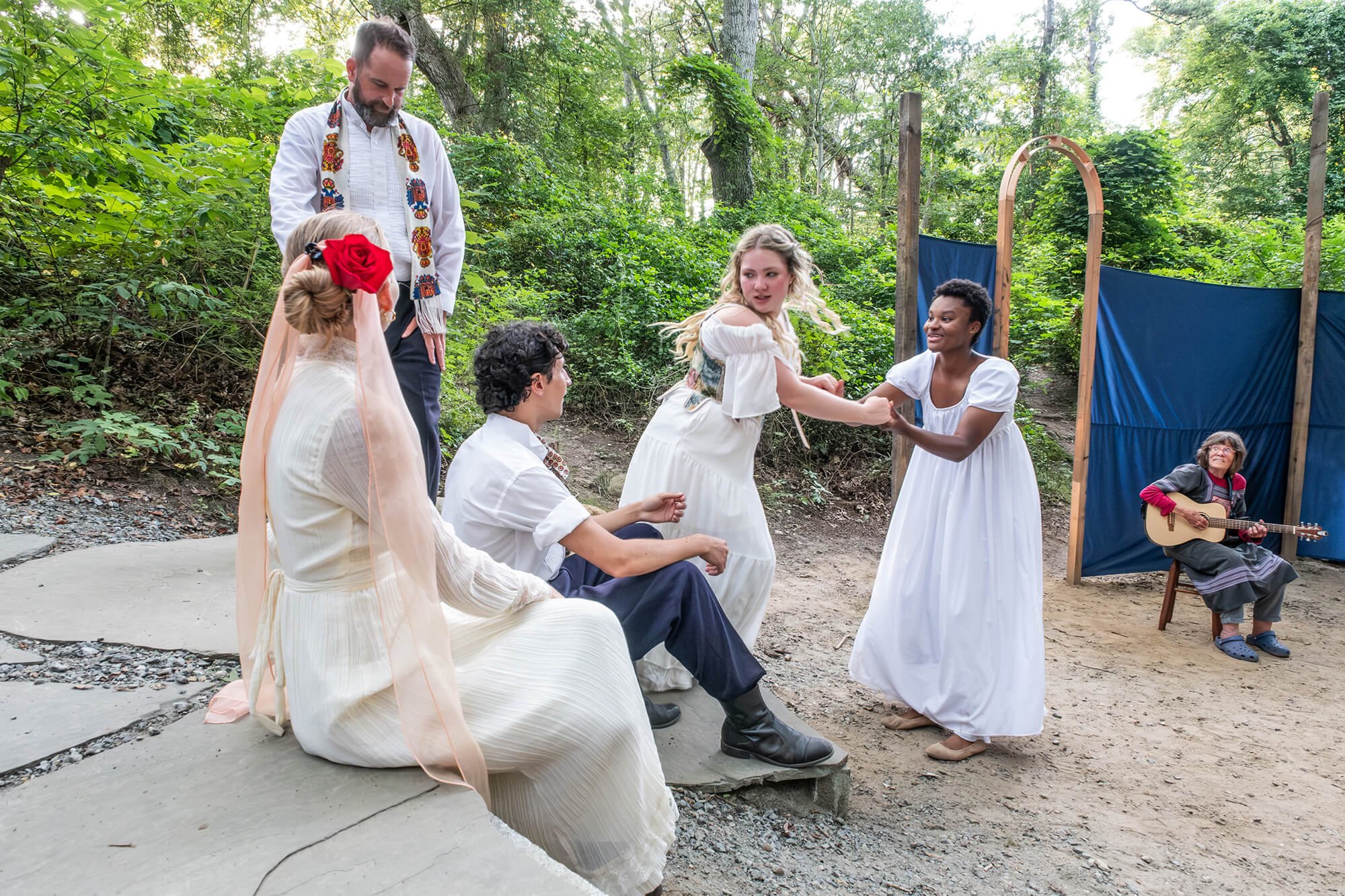Group of people performing a play outdoors in a wooded area. The scene includes individuals in period costumes, and a woman playing guitar.