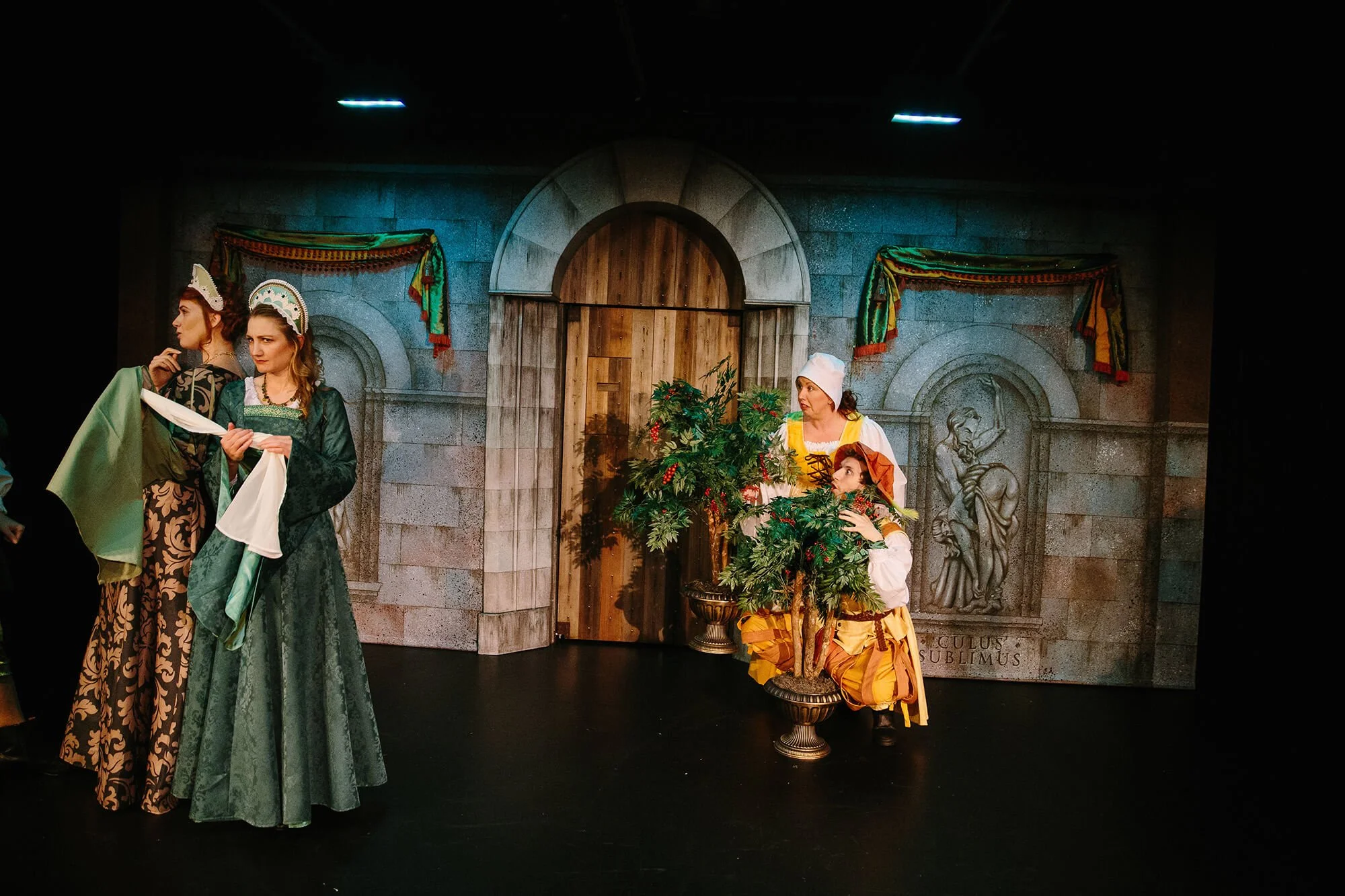 Theater scene with four women in historical costumes, stage set resembling a castle wall with arches and Latin inscriptions, decorative banners, and a potted plant.
