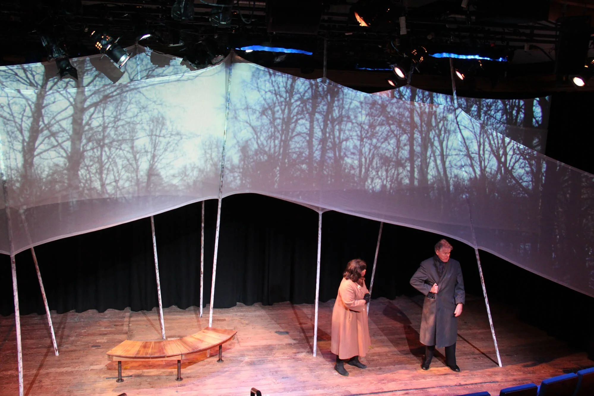 Two people standing on a stage, with one man and one woman, under a projection of leafless trees and a cloudy sky, with a curved fabric or paper canopy overhead.