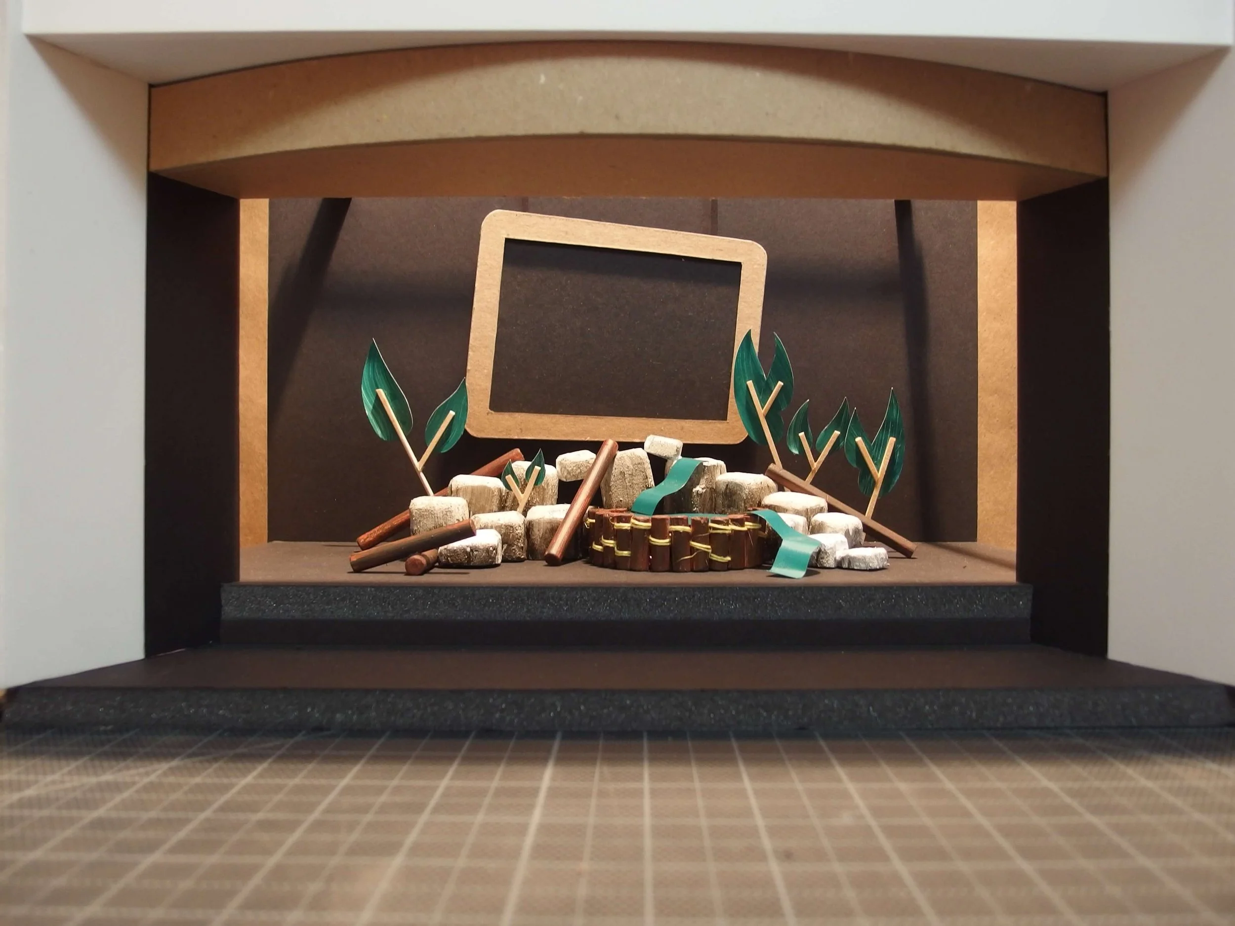 Miniature indoor scene of a classroom or workshop setting, with small trees, a blank chalkboard, and a small stage or platform.