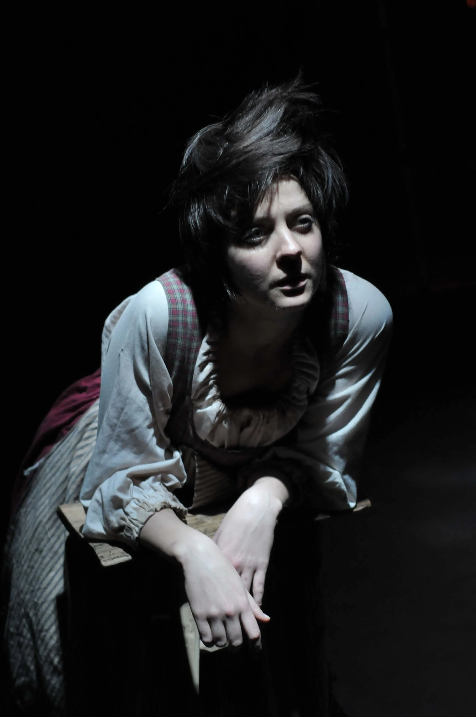 A woman with disheveled hair sitting on a wooden surface, illuminated by a spotlight with a dark background, wearing period costume.