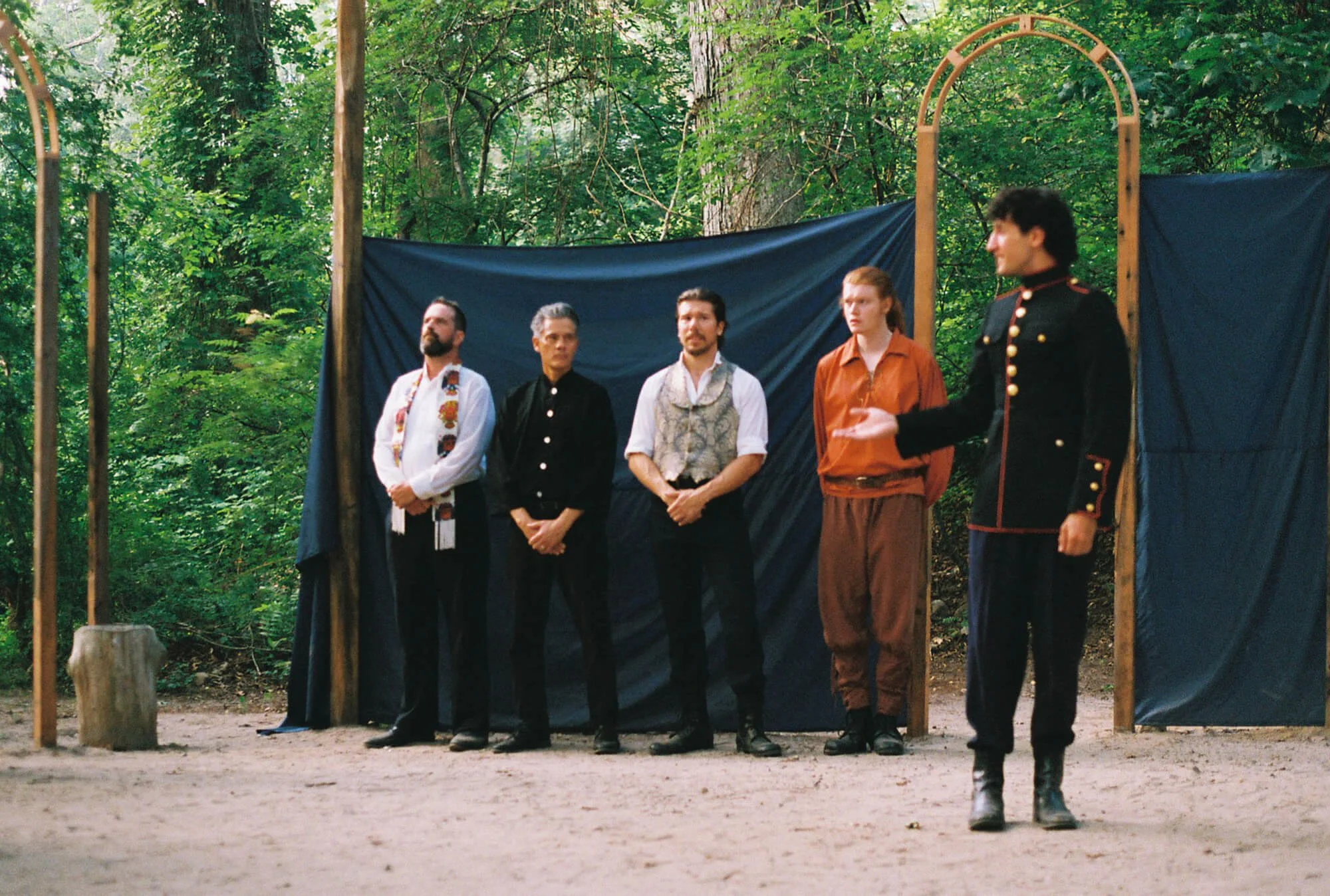 A scene from an outdoor theatrical performance in a wooded area with five actors on stage. Four actors stand side by side, and one actor, dressed in a black military-style uniform with gold buttons, stands apart, gesturing towards the group.