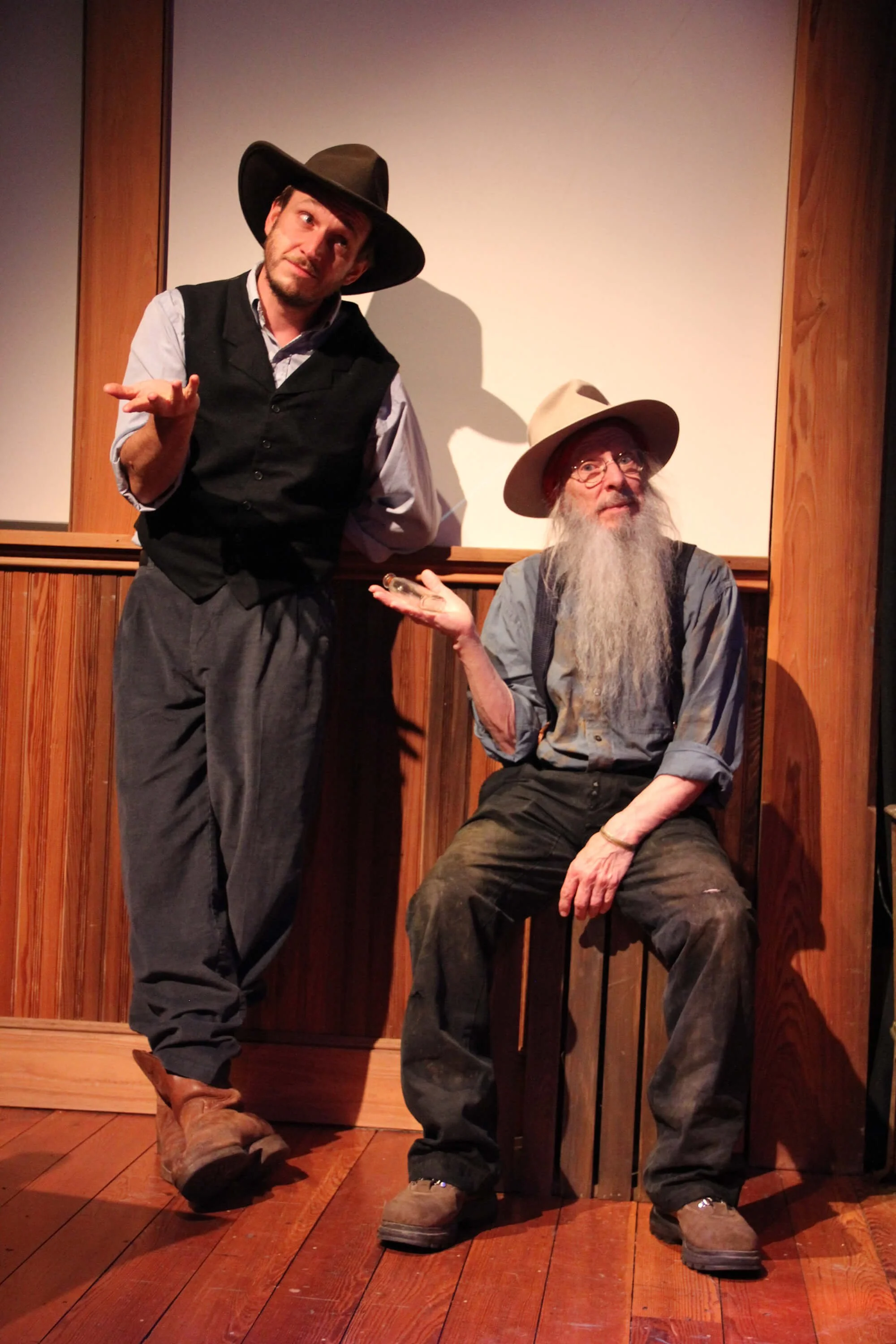 Two men dressed as old-fashioned cowboys or ranchers, one standing and the other sitting, inside a wood-paneled room. The standing man has a hat, vest, and rolled-up sleeves, and is gesturing with his hand. The seated man has a long gray beard, glasses, a wide-brimmed hat, suspenders, and is sitting on a wooden box.