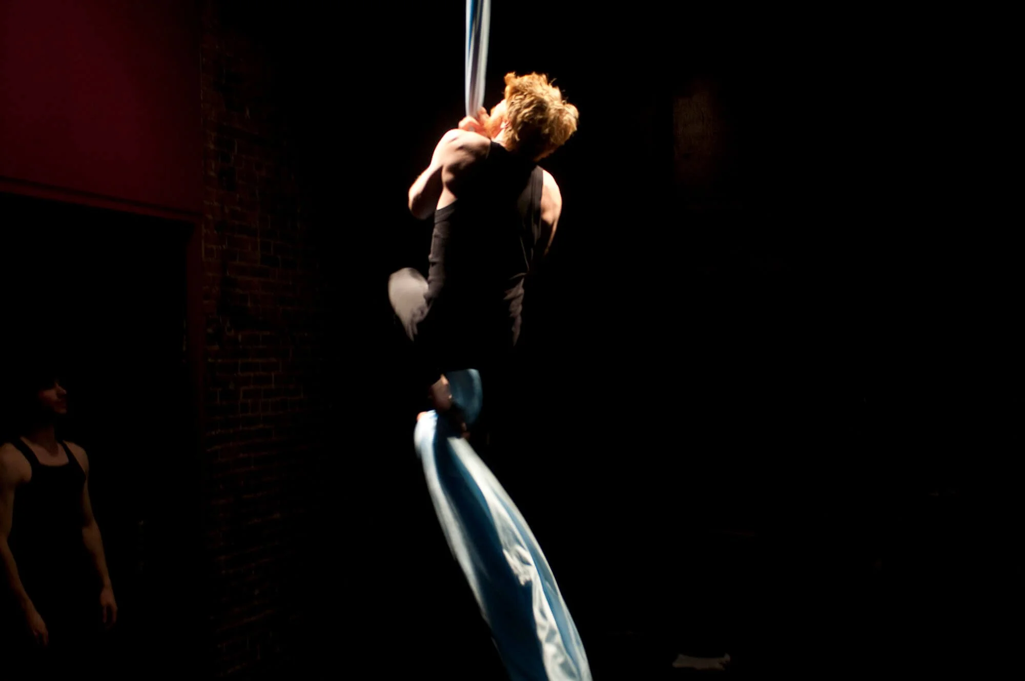 Performer swinging on aerial silk in a dark theater, with a person observing in the background.
