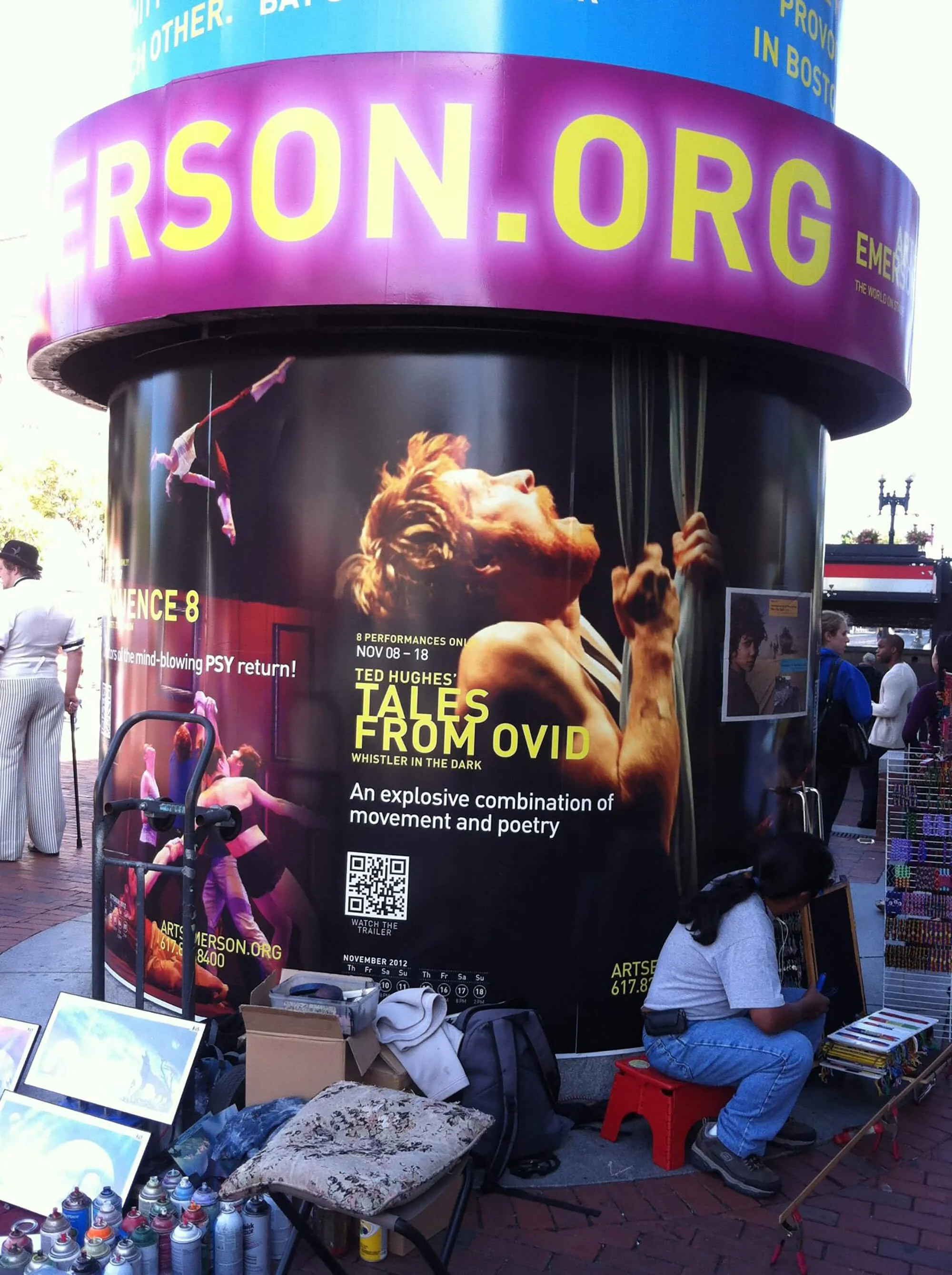 Large advertising display for Emerson.org featuring a promotional poster for TED Hughes' 'Tales from Ovid' performance, with images of performers and a QR code, set on a cylindrical structure surrounded by street vendors and pedestrians.