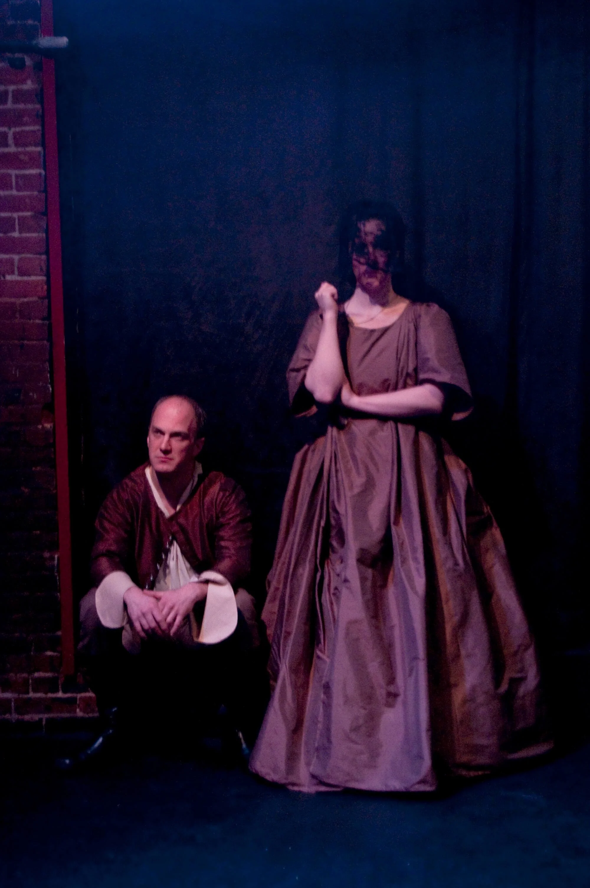 Two actors on stage in historical costumes, one squatting and the other standing, with a dark background and a brick wall on the side.