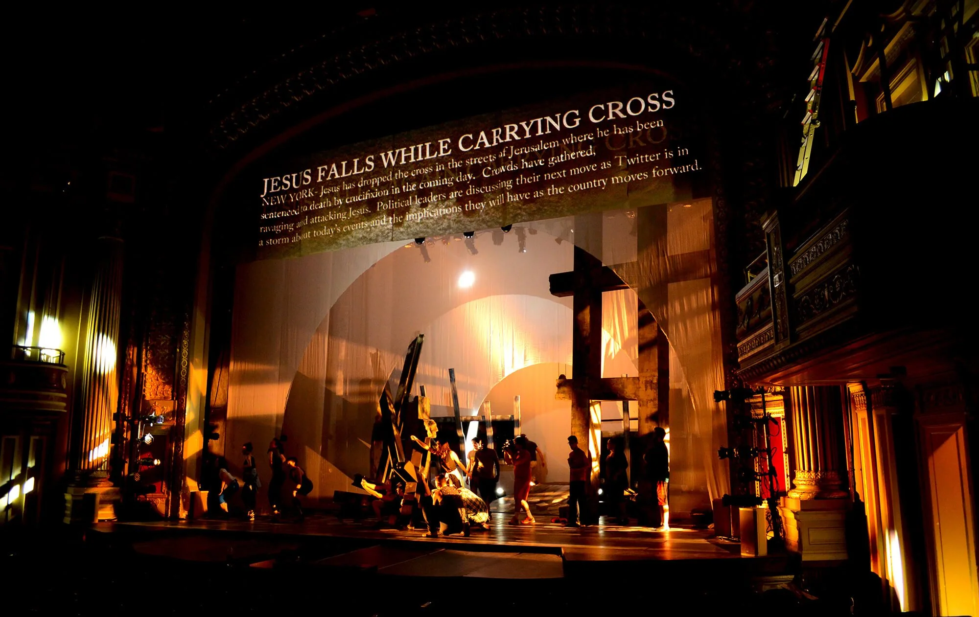 Stage set with a religious-themed scene, featuring silhouettes of people, a large cross, and a backdrop with a projected headline about Jesus falling while carrying the cross.