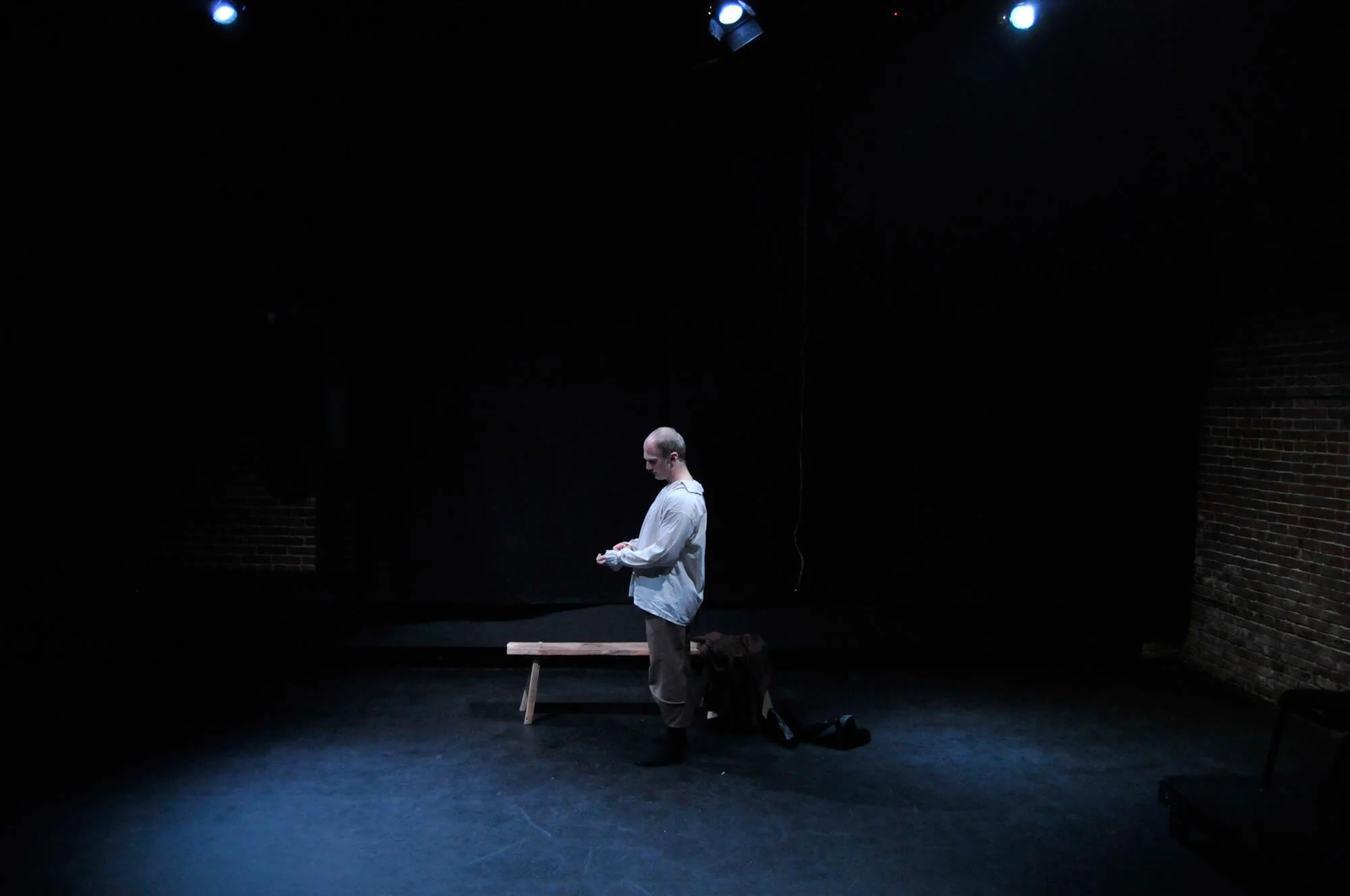 A person standing alone on a dark stage under blue spotlights, next to a wooden bench and a black backpack, with brick walls on either side.