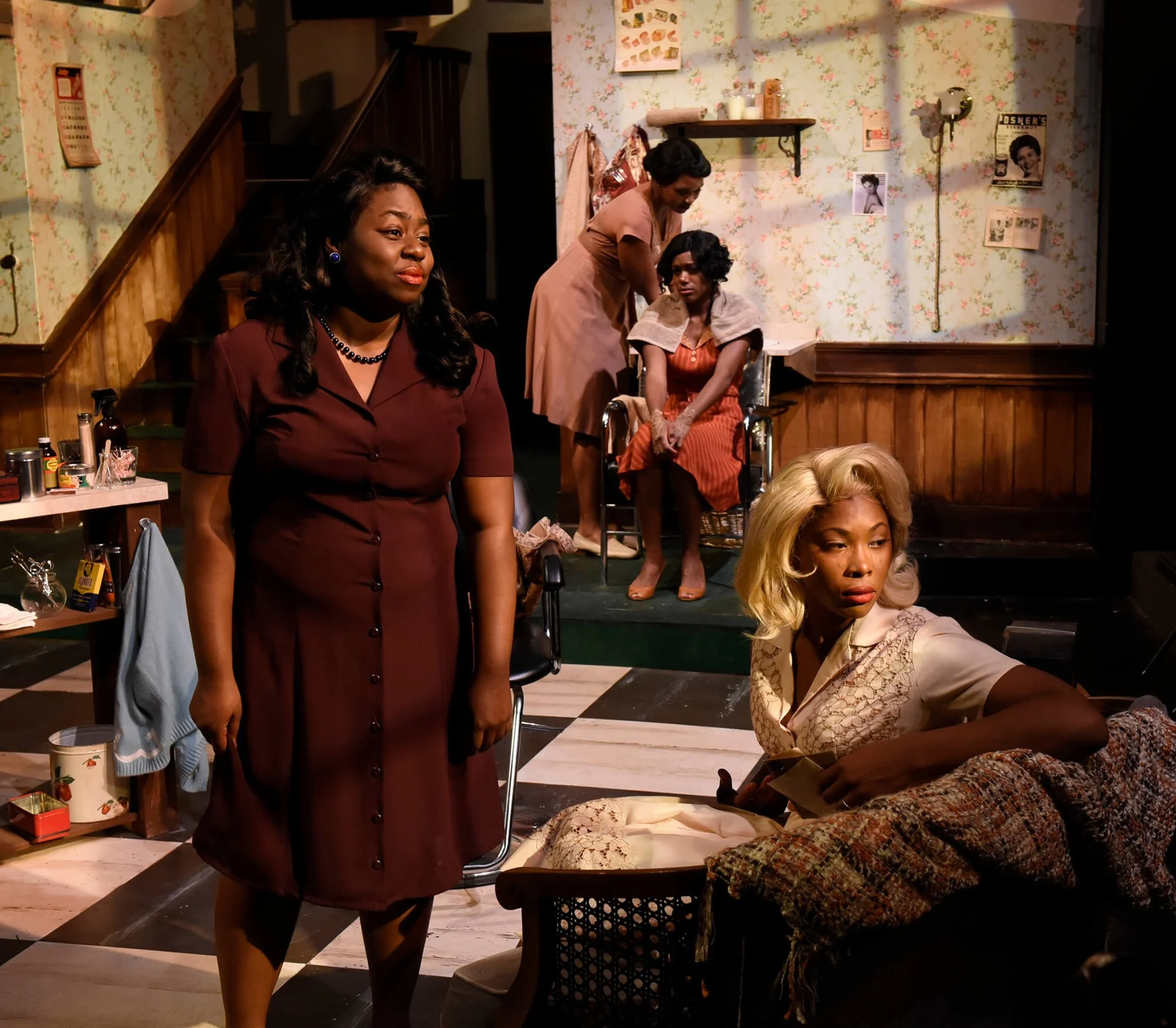 Women in a retro-style living room, with one woman sitting on the couch and another standing beside her, while two women are in the background, one sitting in a wheelchair and the other standing behind her, all engaged in an emotionally charged scene