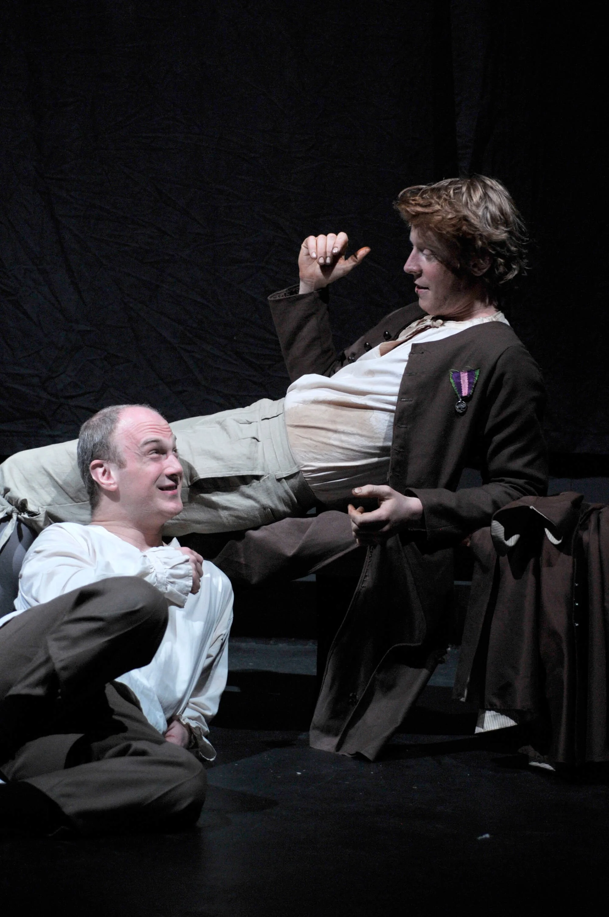 Two performers on stage, one sitting on the floor and the other reclined, engaged in a theatrical scene with a black backdrop.