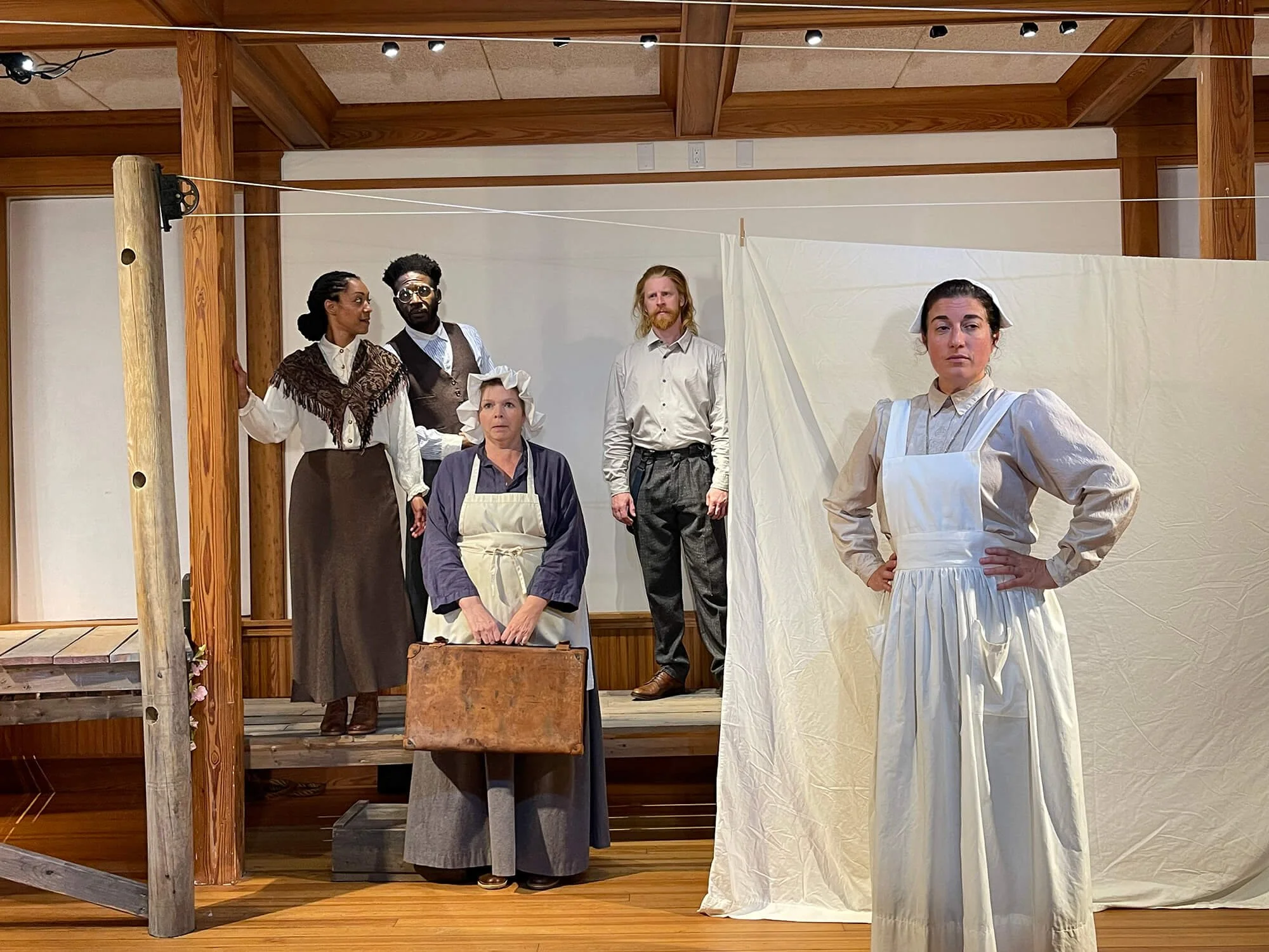 The image shows five actors on a stage dressed in costumes from a historical play, with some standing on floor level and others on a raised platform. The setting appears to be a rustic, wooden interior, possibly a theater or a stage set.
