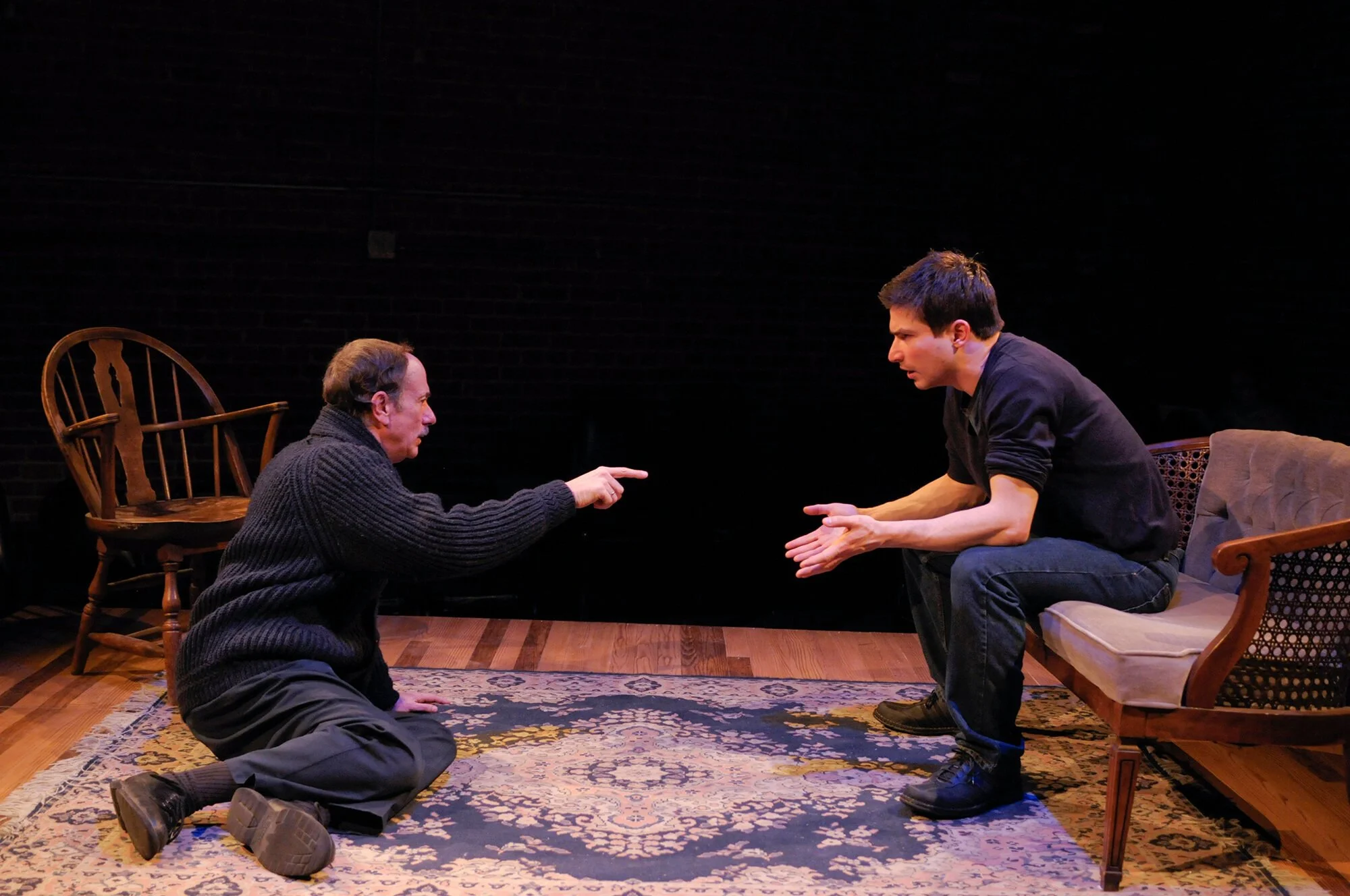Two men in a theatrical scene, one sitting on a couch with an intense expression, the other kneeling on the floor pointing and speaking passionately, both engaged in a dramatic conversation on a stage with a dark background.