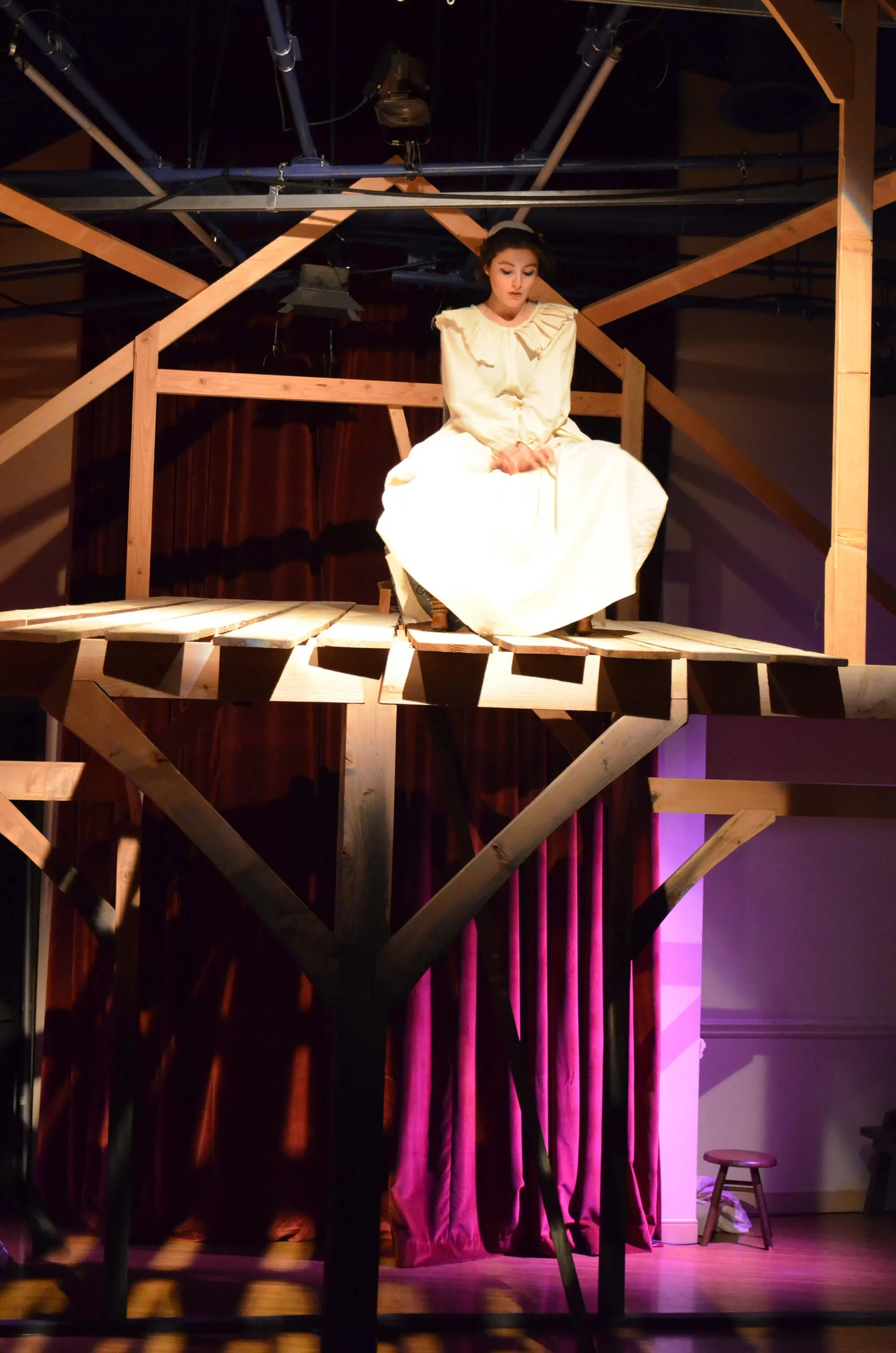 A woman in a white dress is sitting on a wooden platform stage with a wooden structure around her in a theater. She has dark hair and appears to be looking down.