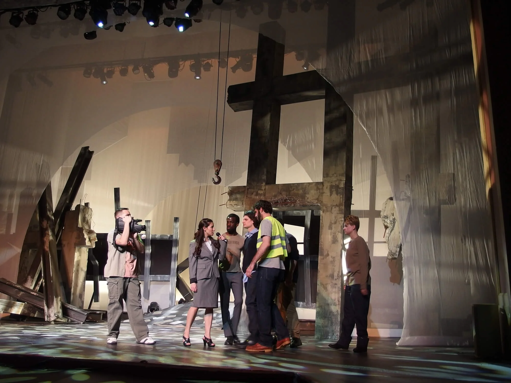 People on stage, with one person interviewing another, in front of a set that looks like a destroyed building with debris and a large cross, during a theatrical or film production.