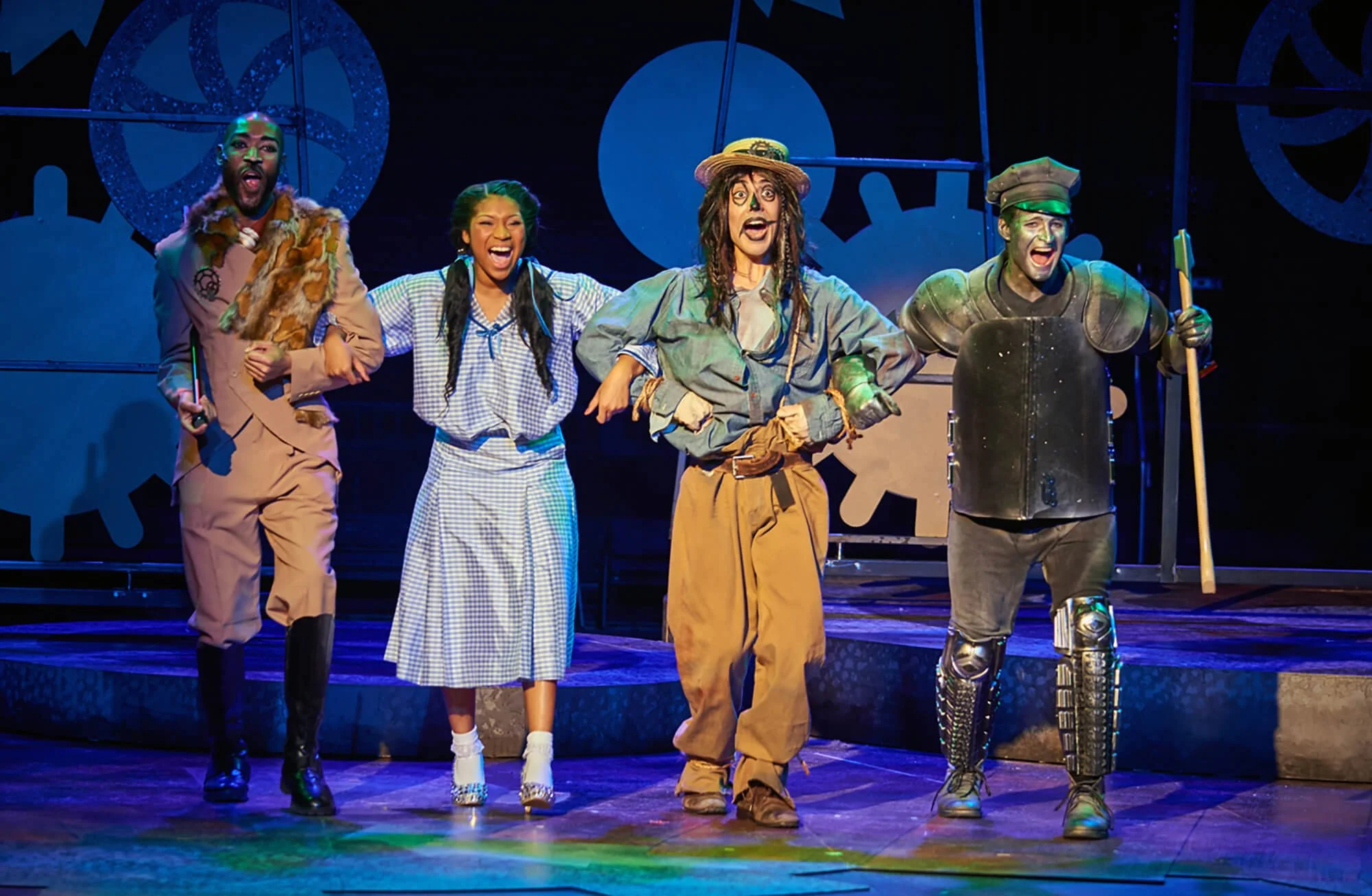 The cast of "The Wizard of Oz" musical perform on stage, dressed in costumes representing the characters, with gears and cogs in the background.