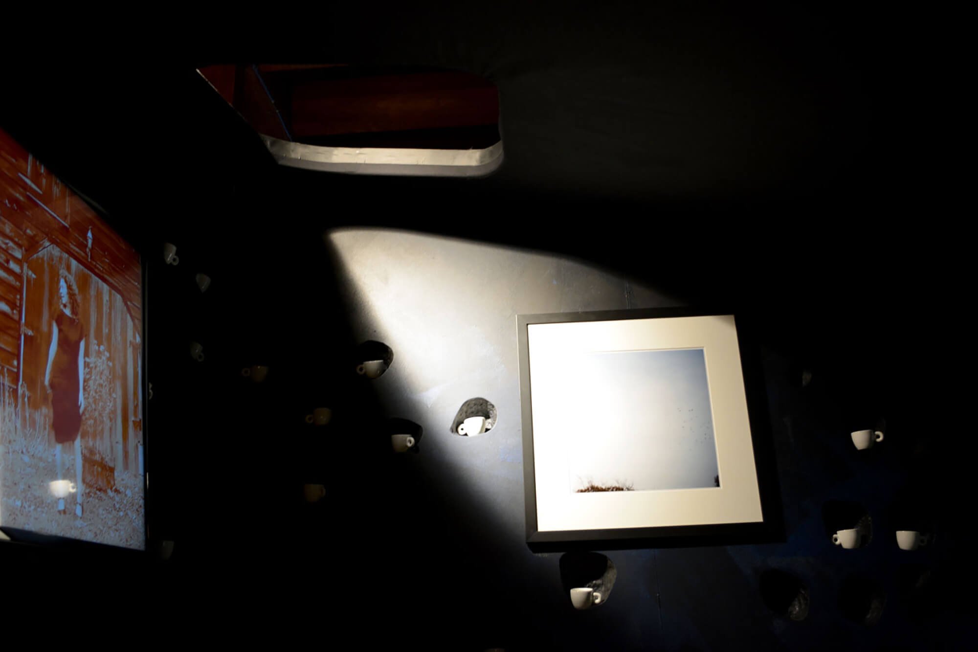 A top-down view of a display with a photograph, a framed picture, and several small cups arranged on a dark surface with spot lighting.