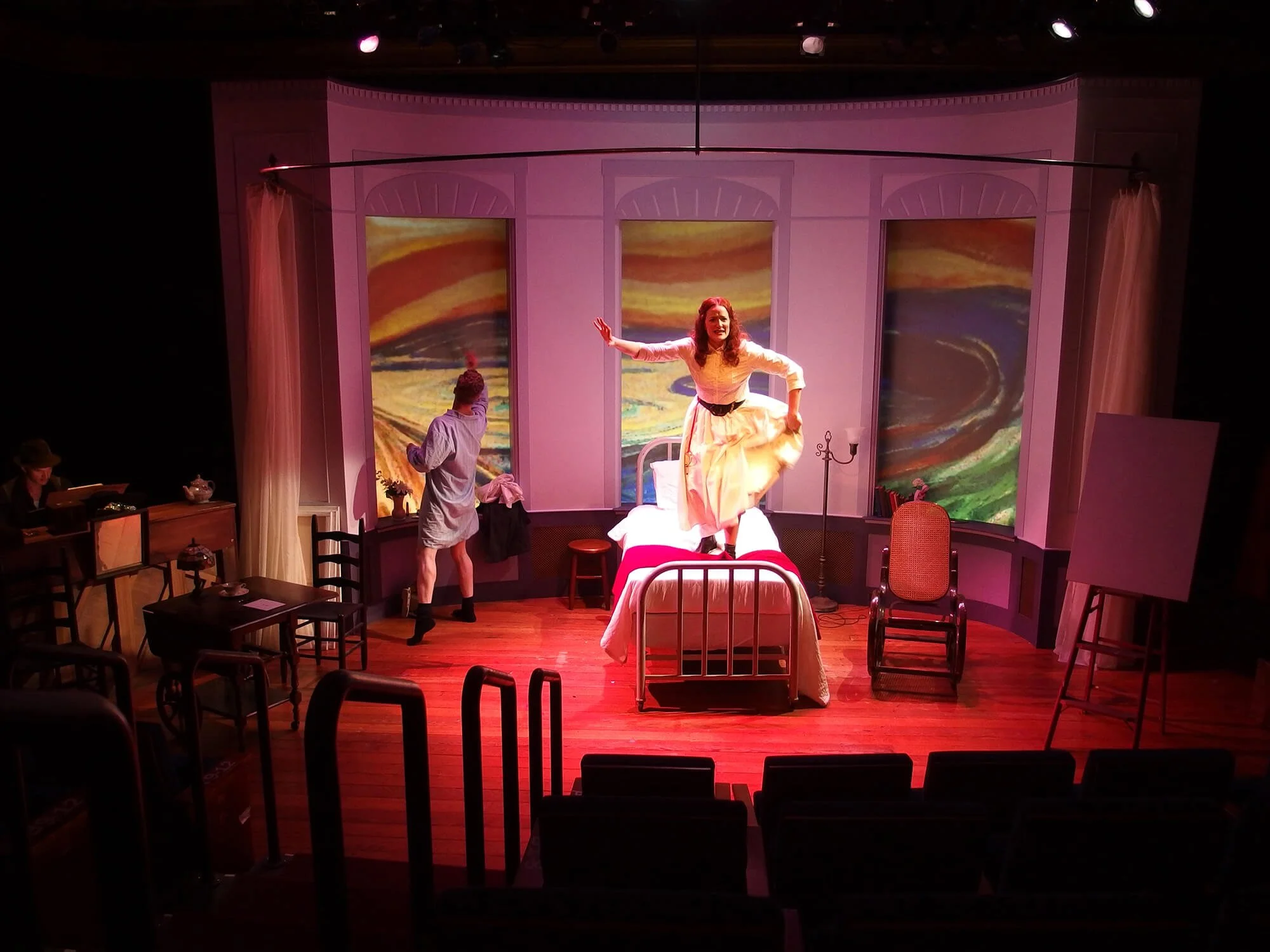 The stage set for a theatrical performance features a woman in a yellow dress standing on a bed, with her arms outstretched, and back-up actor in a nightshirt and socks. The backdrop displays colorful, swirling artwork. There are props, chairs, and a