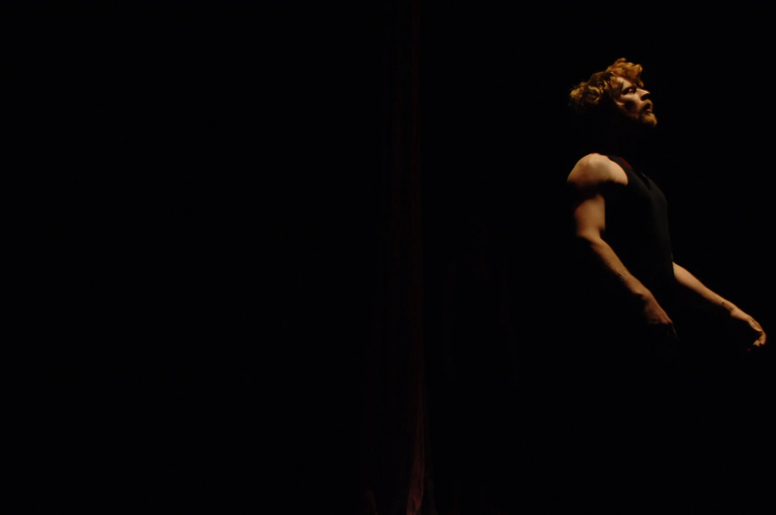 A person with curly hair and a beard standing on stage in a dark setting, illuminated from the side.
