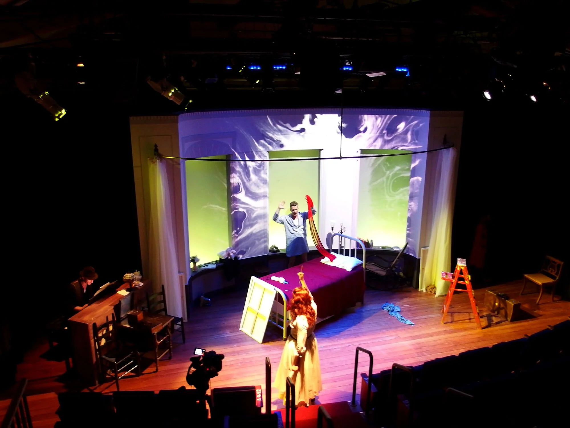 A stage set depicts a scene in a bedroom with a person standing in front of the bed and another person in a long dress. The stage has a backdrop with swirling patterns, furniture including chairs and a table, and props like a ladder and a picture fra