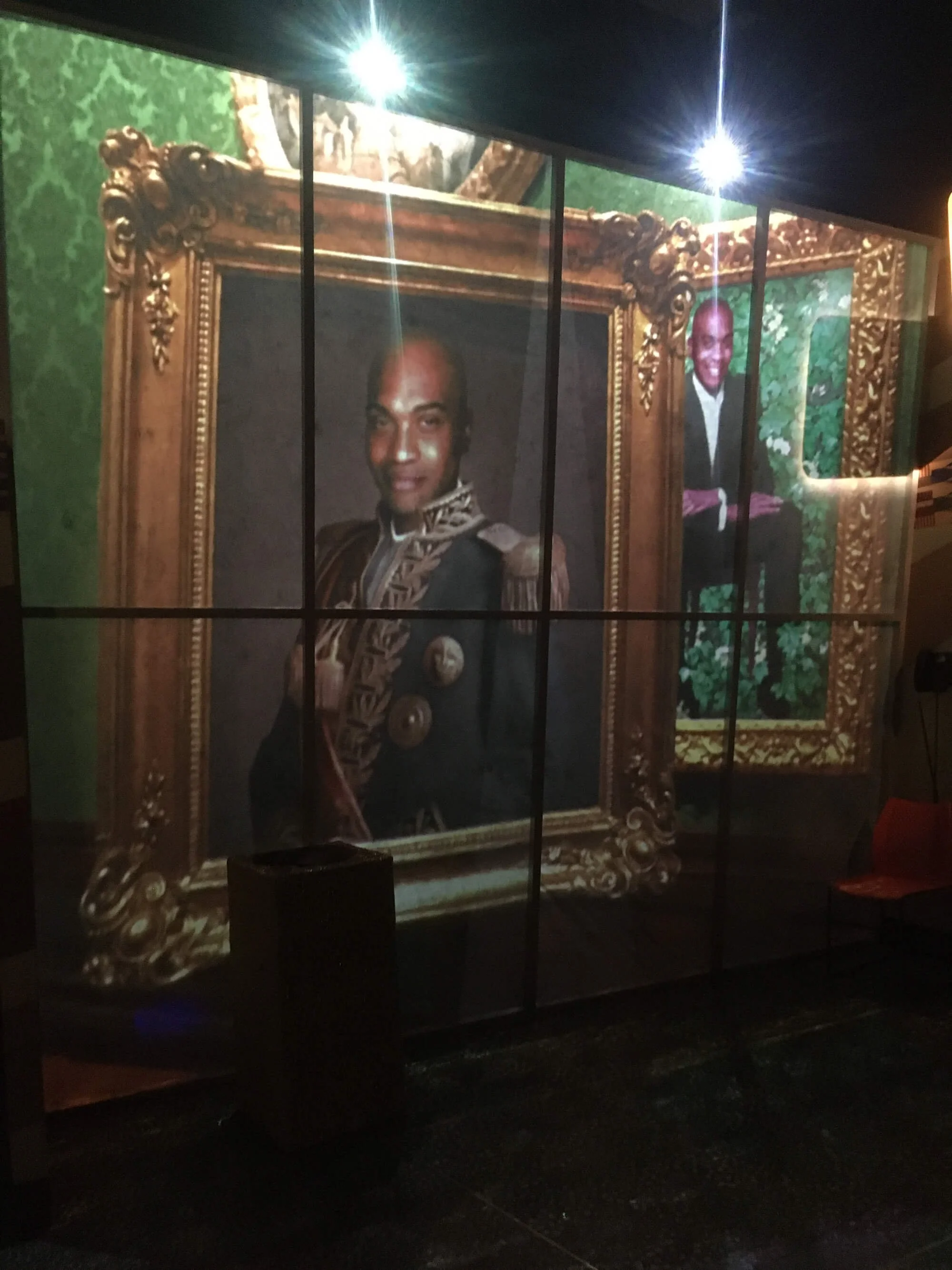 A large screen showing artwork featuring two men, one in historical attire with medals, and the other in modern clothing, each framed by ornate golden frames, set against a green background with foliage.