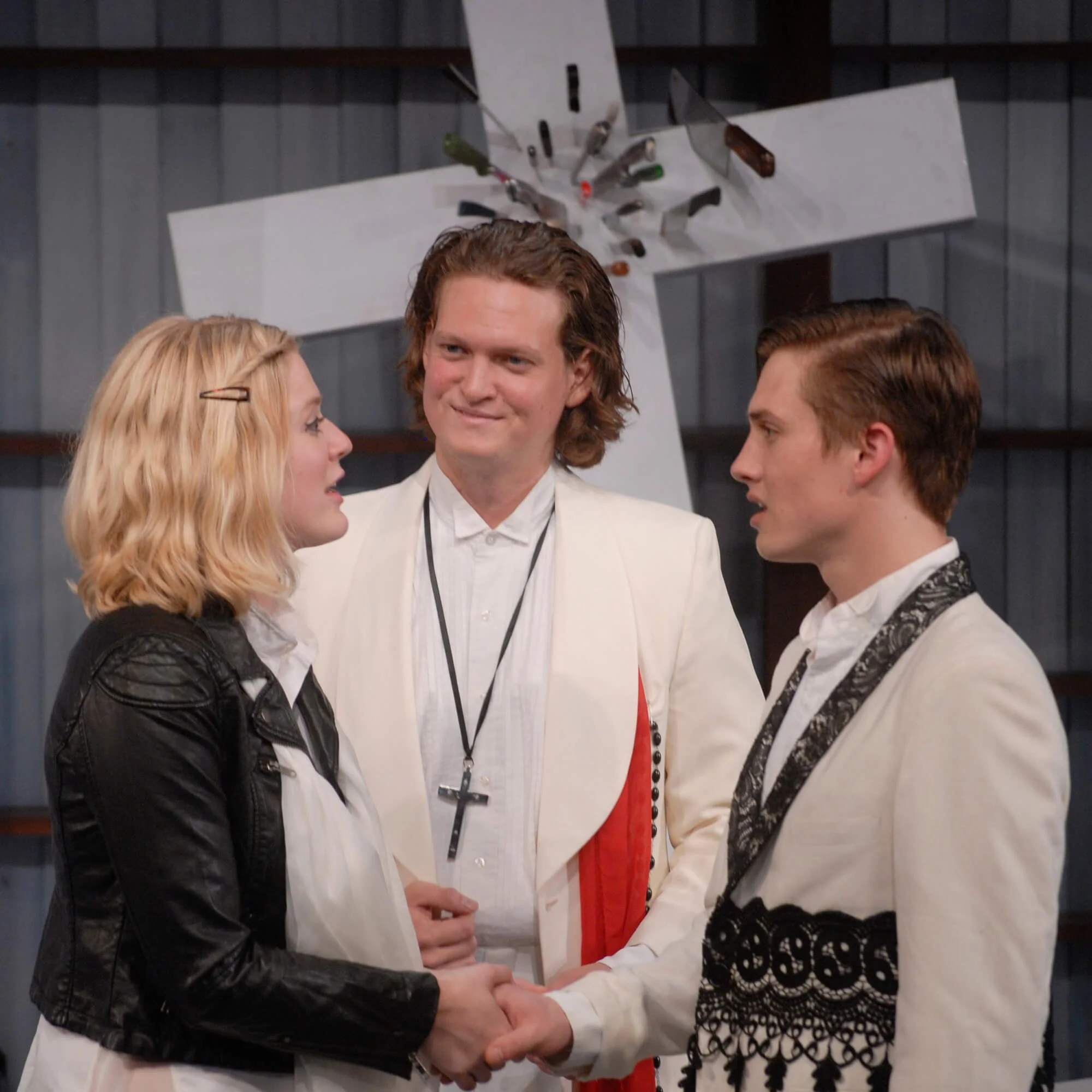 Three young adults, two men and one woman, engaged in a handshake during a formal event, with a large metal cross with knives attached hanging behind them.