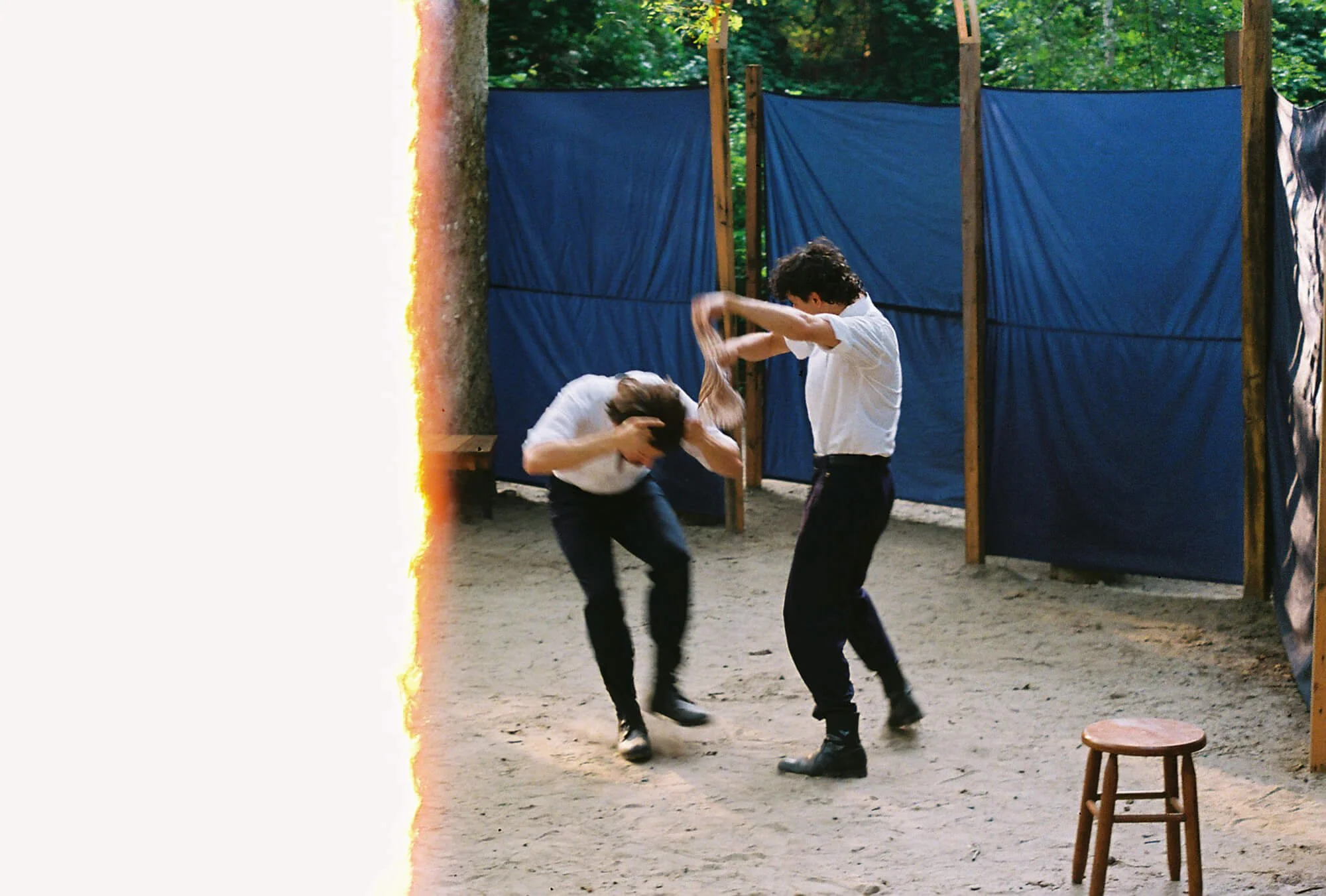 Two boys in white shirts and dark pants fighting or playing aggressively in a wooded outdoor area with blue fabric screens behind them and a small wooden stool nearby.