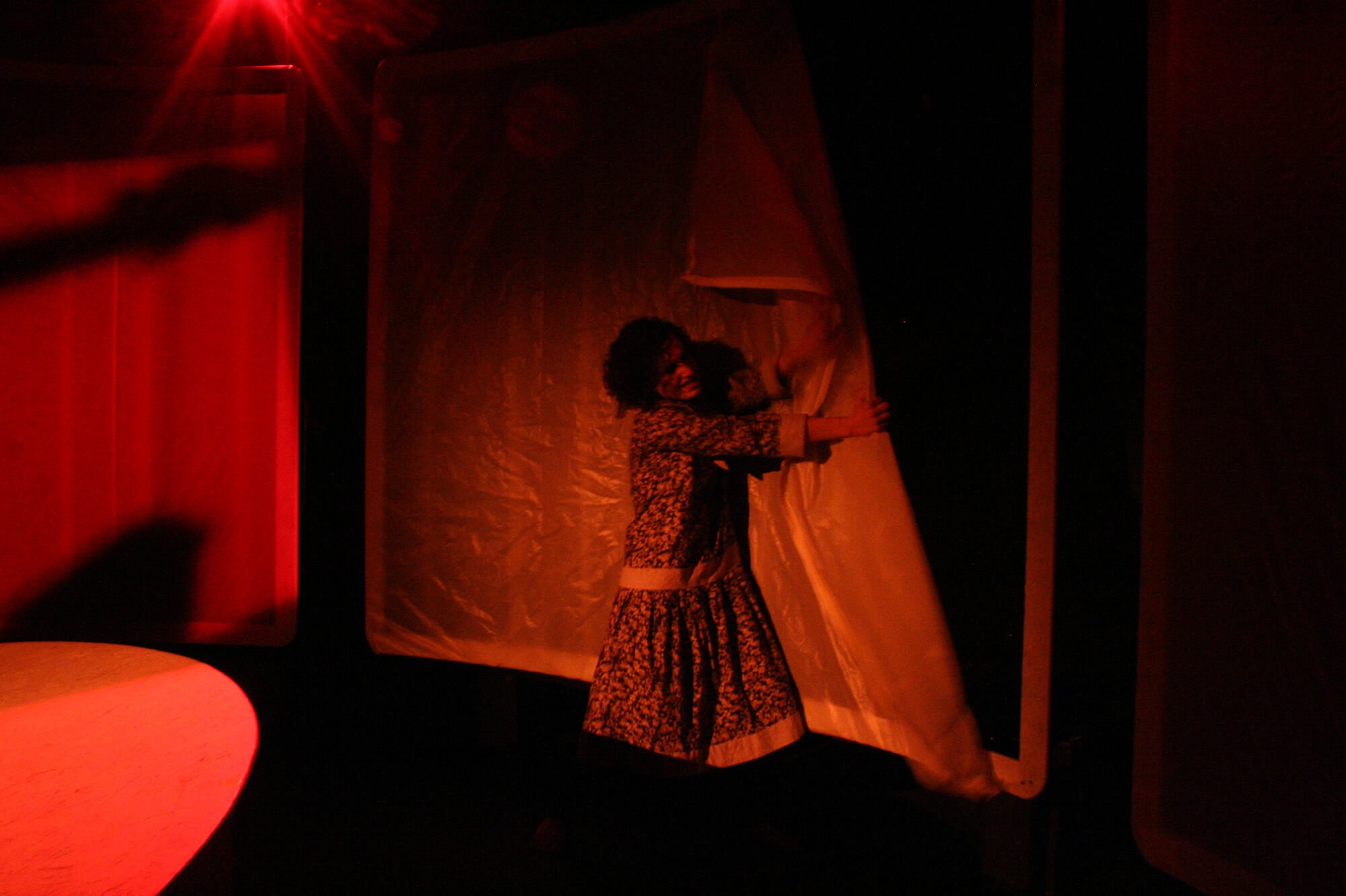 A woman with curly hair wearing a patterned dress standing on a stage, pulling back a curtain with a smile, illuminated by red lighting.