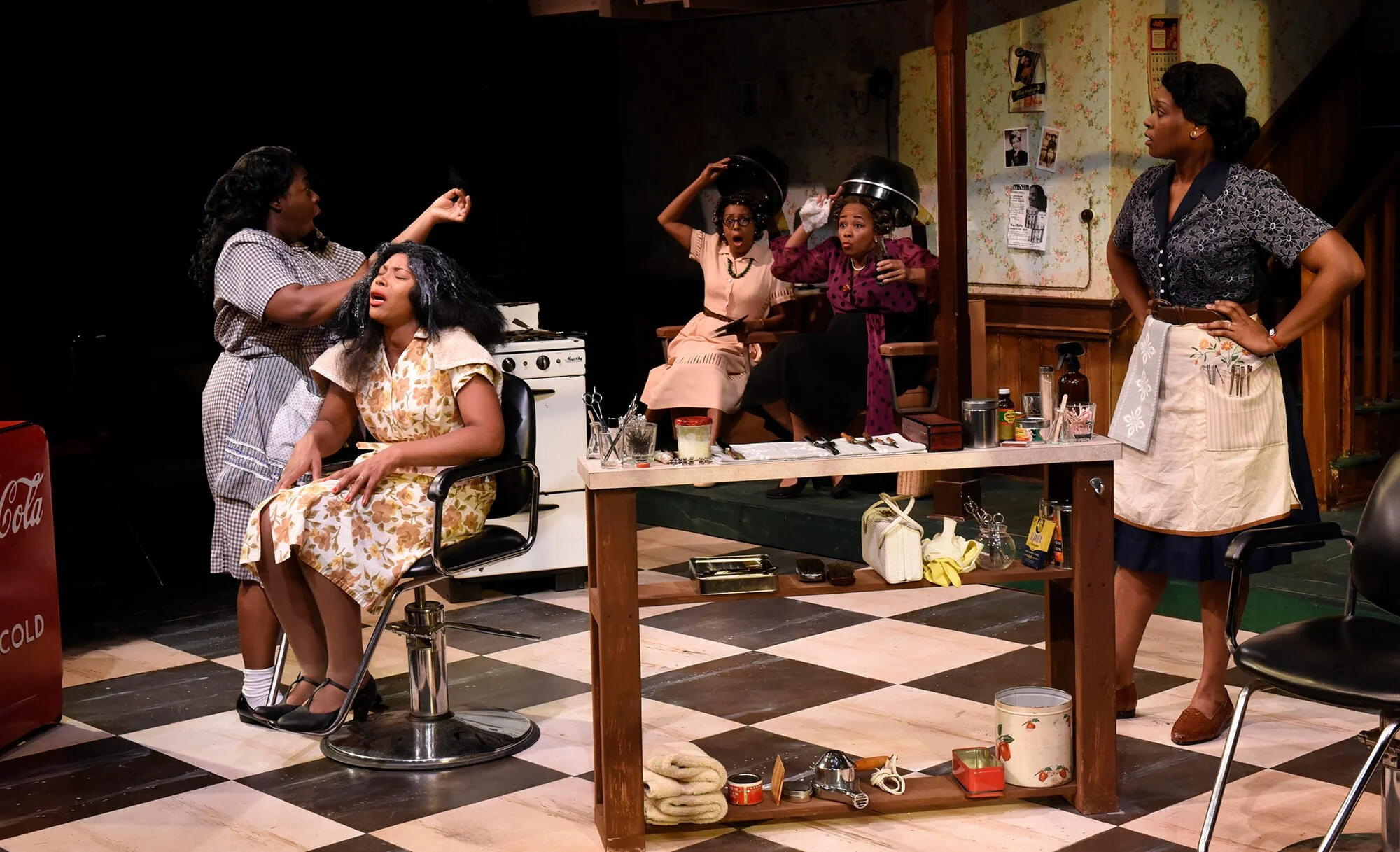 Scene in a hair salon with five women, one sitting in a stylist's chair being attended to, three in the background making shocked expressions with hair dye shields on their heads, and one standing with hands on hips watching the scene.