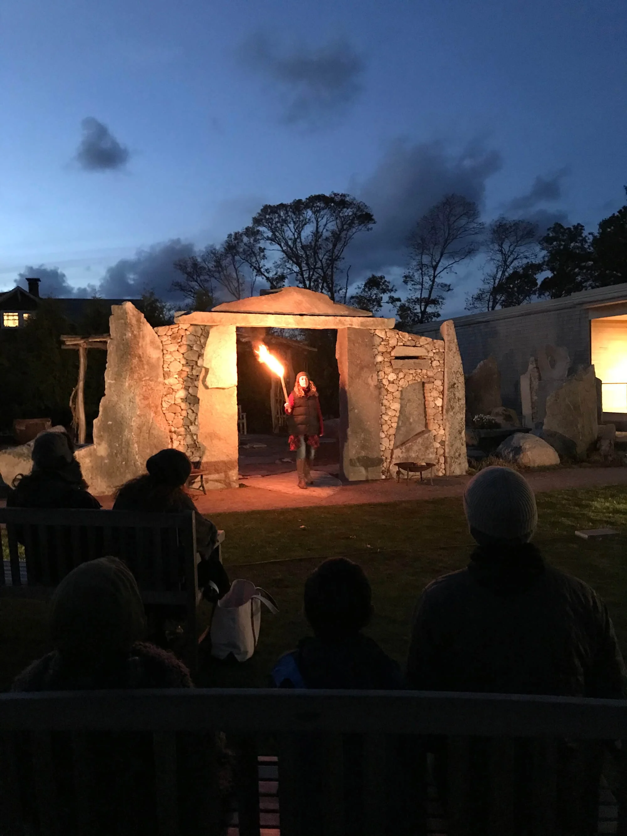 Person standing in front of stone structure at dusk, holding a flaming torch, with seated audience watching.