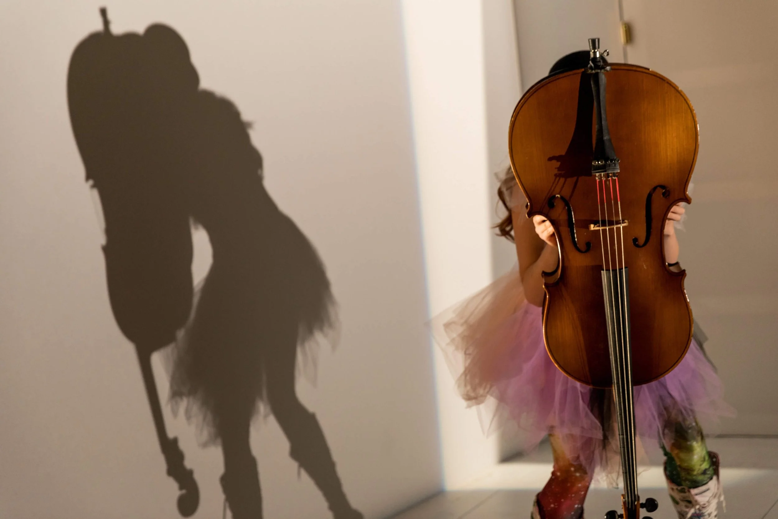A young girl in a pink tutu and patterned leggings is holding a large wooden cello, obscuring her face and upper body. Her shadow is cast on the wall behind her.