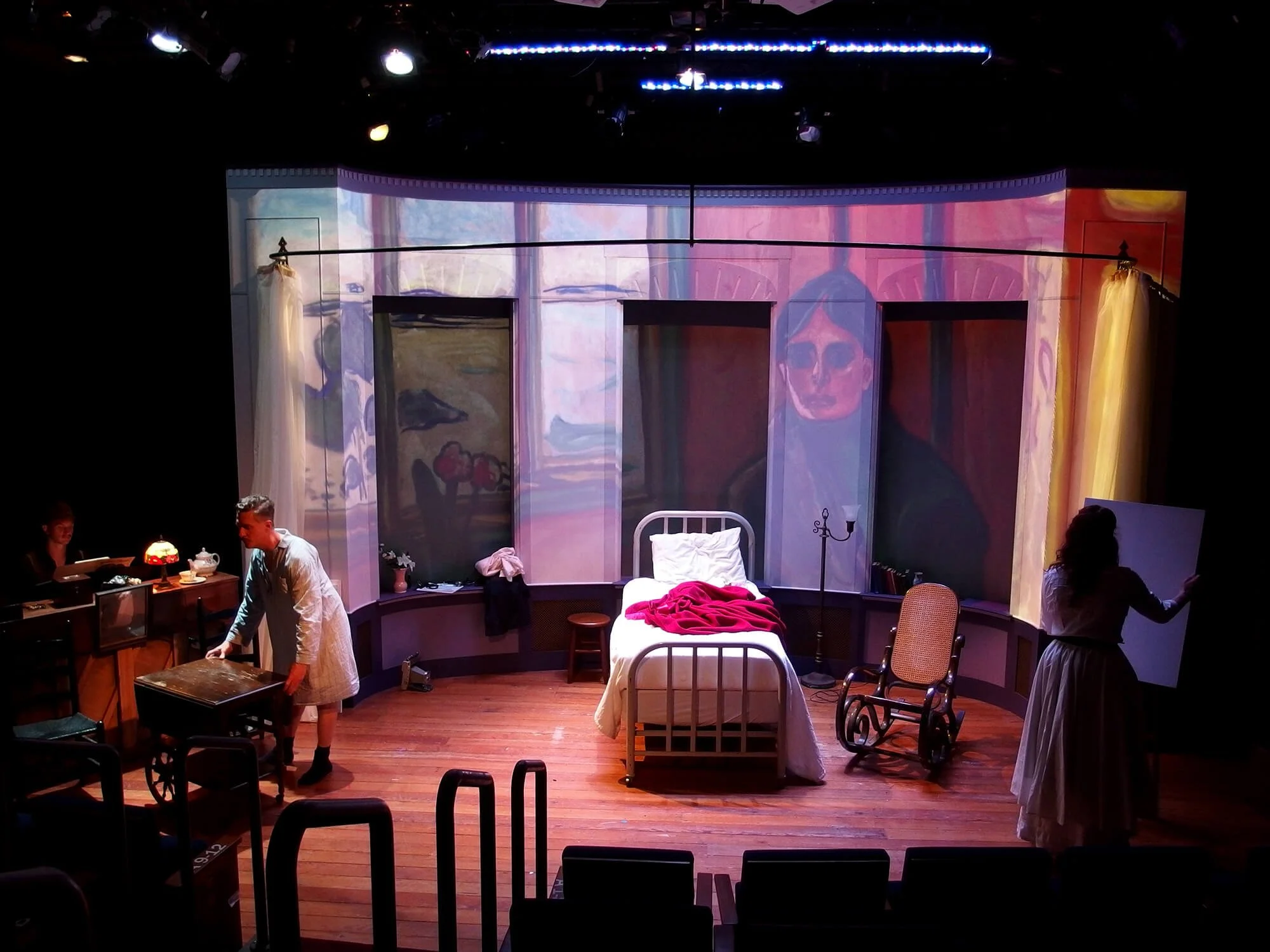 Stage set designed to resemble a bedroom with a painted backdrop of a woman wearing sunglasses. The stage includes a white bed with a red blanket, a wooden stool, a rocking chair, and a woman writing on a whiteboard. Stage lights illuminate the scene