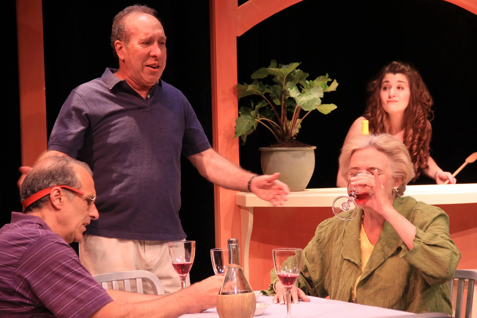 Four people at a dinner party, with one woman drinking wine, a man standing and speaking, and another man sitting at the table. There is a woman in the background behind a counter, and a potted plant near the counter.