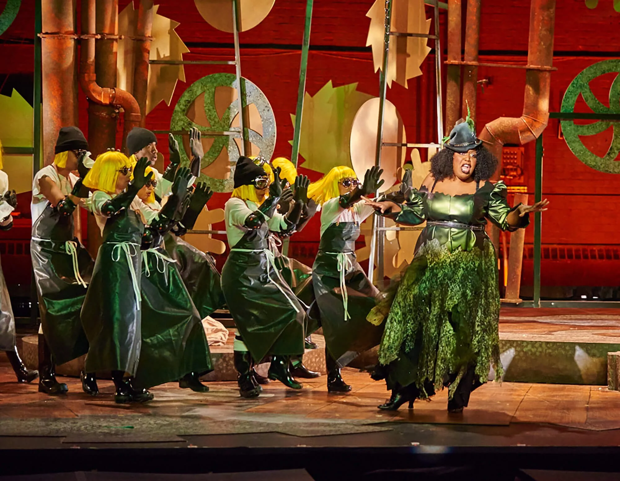 Stage performance with a woman in a green and black costume and a large black hat, surrounded by performers with yellow wigs, sunglasses, black gloves, and green outfits, against a theatrical set background with industrial pipes and large animated co