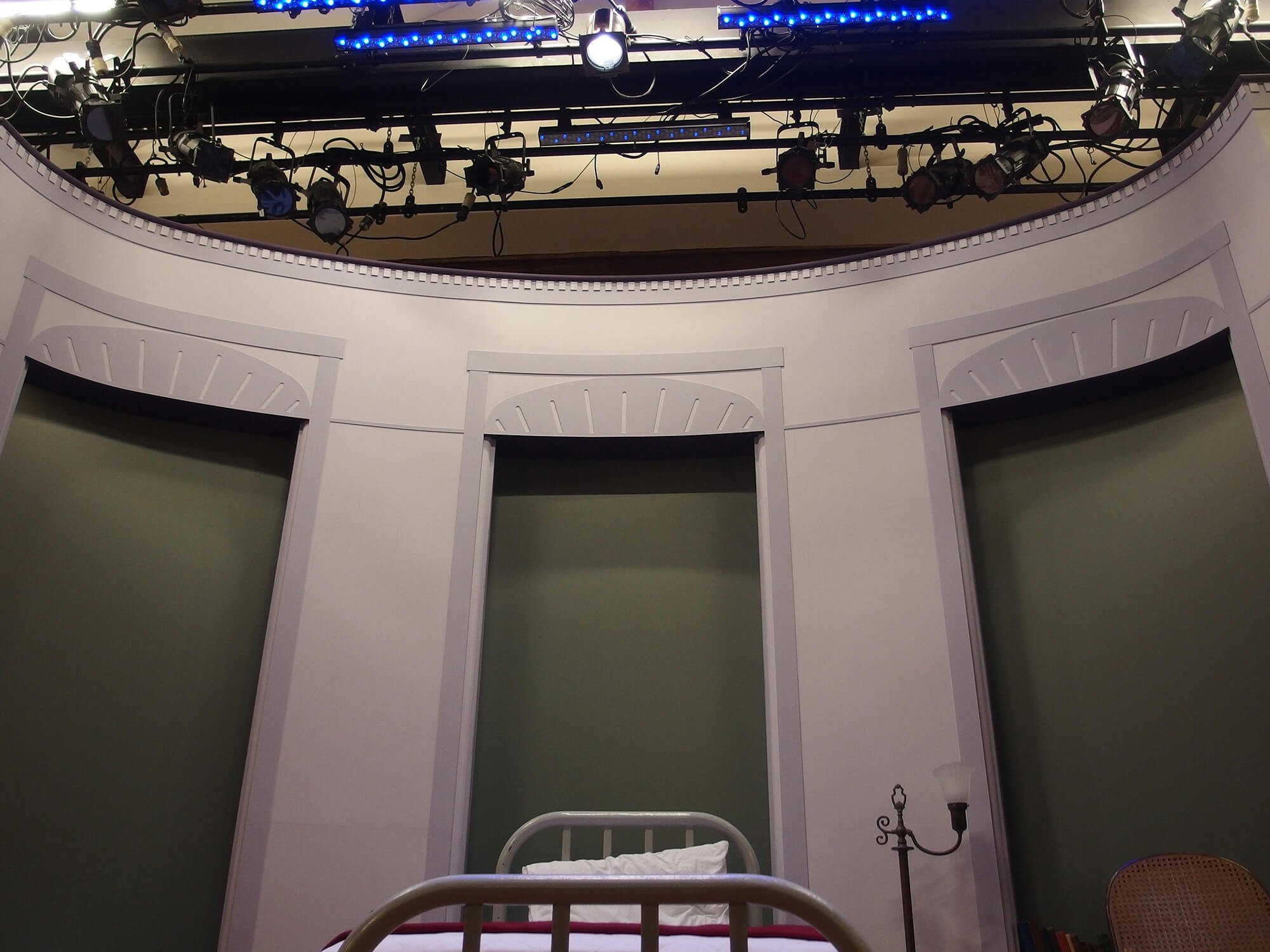 Set of three green panels with white decorative trim, a white bed frame with a pillow and blankets, a small wooden side table, a black metal stand with a lantern, and a rattan chair, against a curved white wall with shadows and lighting equipment ove