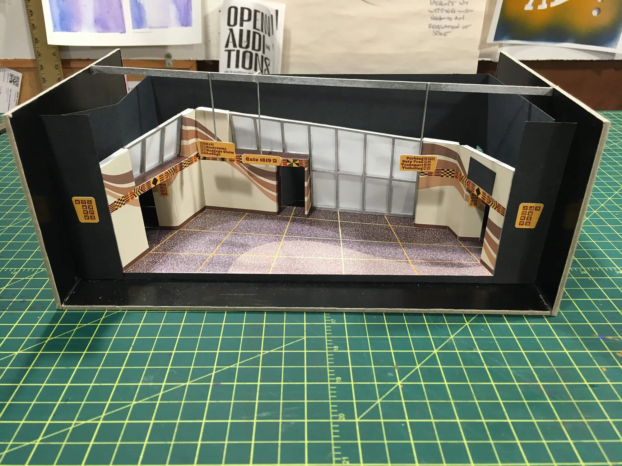 A detailed miniature model of an airport terminal layout with gates, signage, and interior decor, placed on a green cutting mat.
