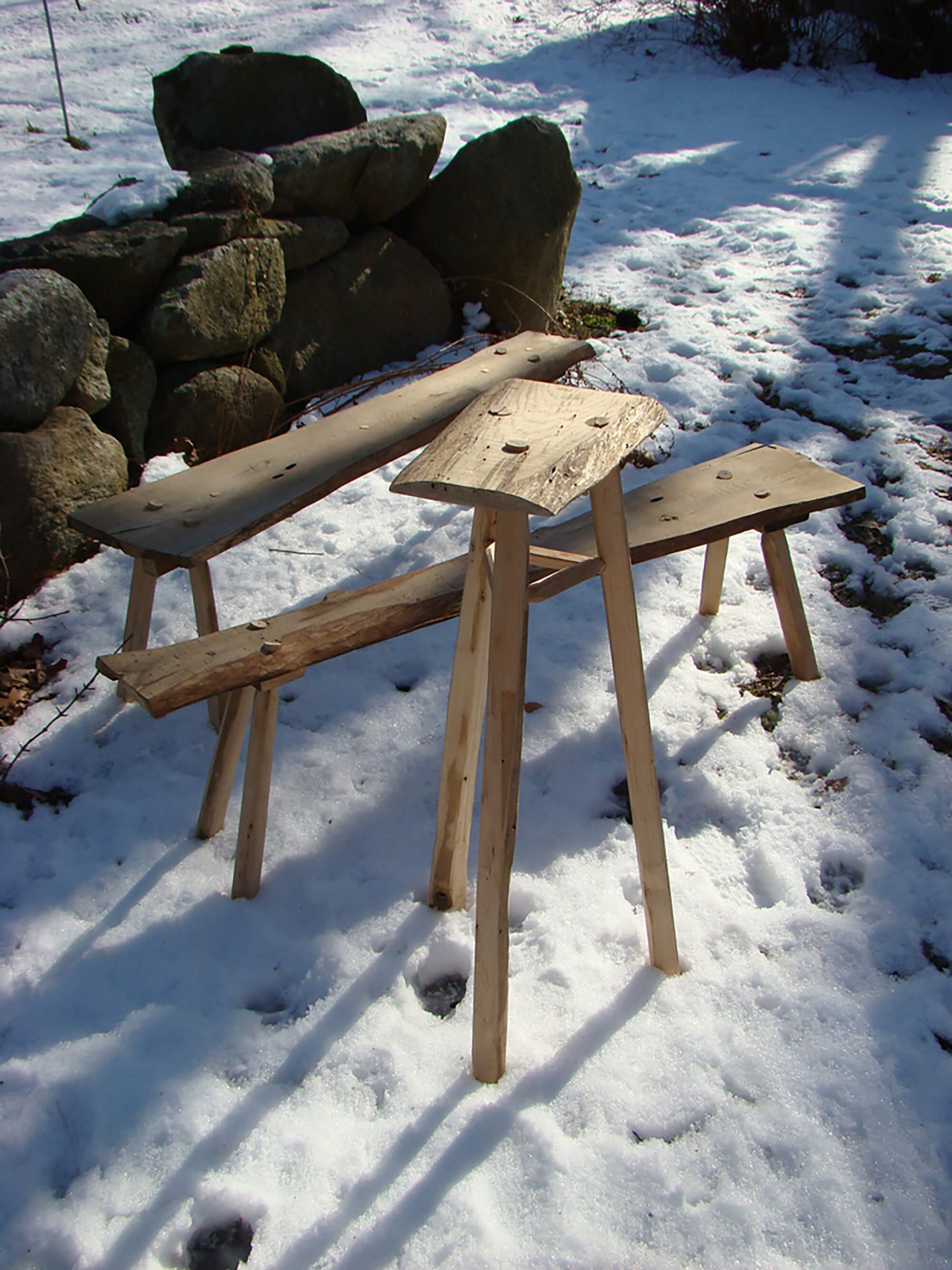 Three handmade wooden stools set on snow-covered ground outdoors, with a stone wall in the background.
