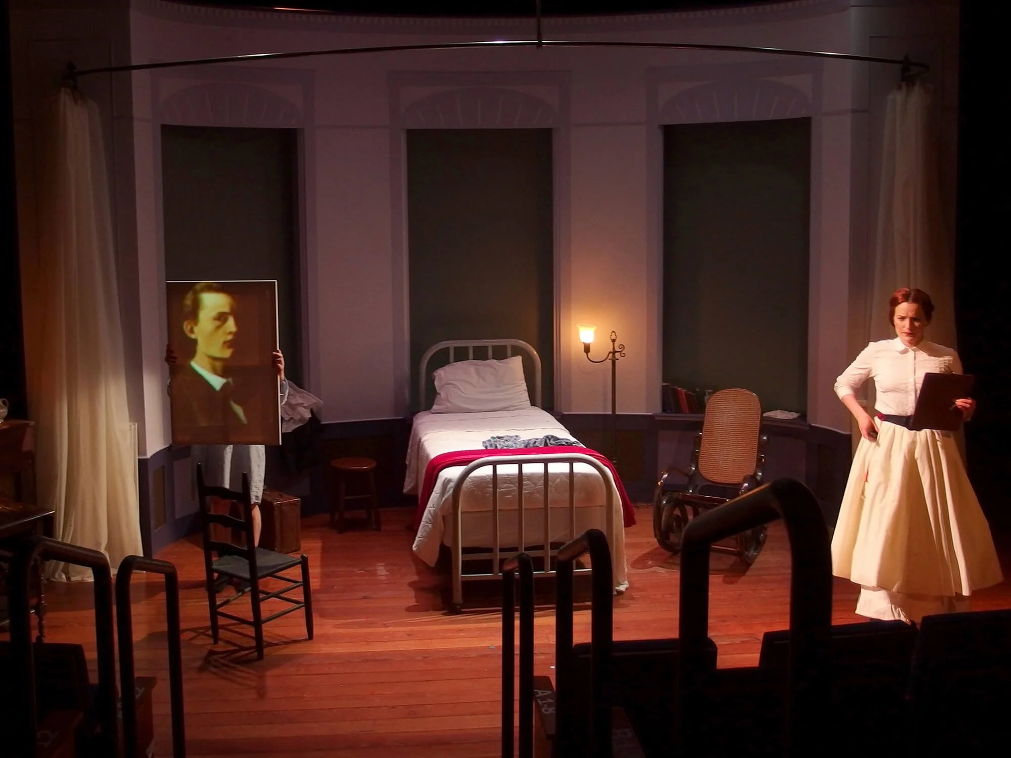 Stage set depicting a bedroom scene with a white bed, a woman in period costume holding a script, a portrait frame, a small table with a lamp, chairs, and a bookcase.
