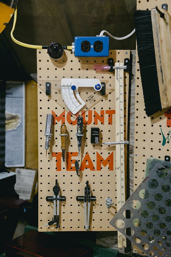 A pegboard with the words "MOUNT TEAM" in orange, holding various tools such as screwdrivers, compasses, and a protractor, with a blue power strip and other miscellaneous workshop items.