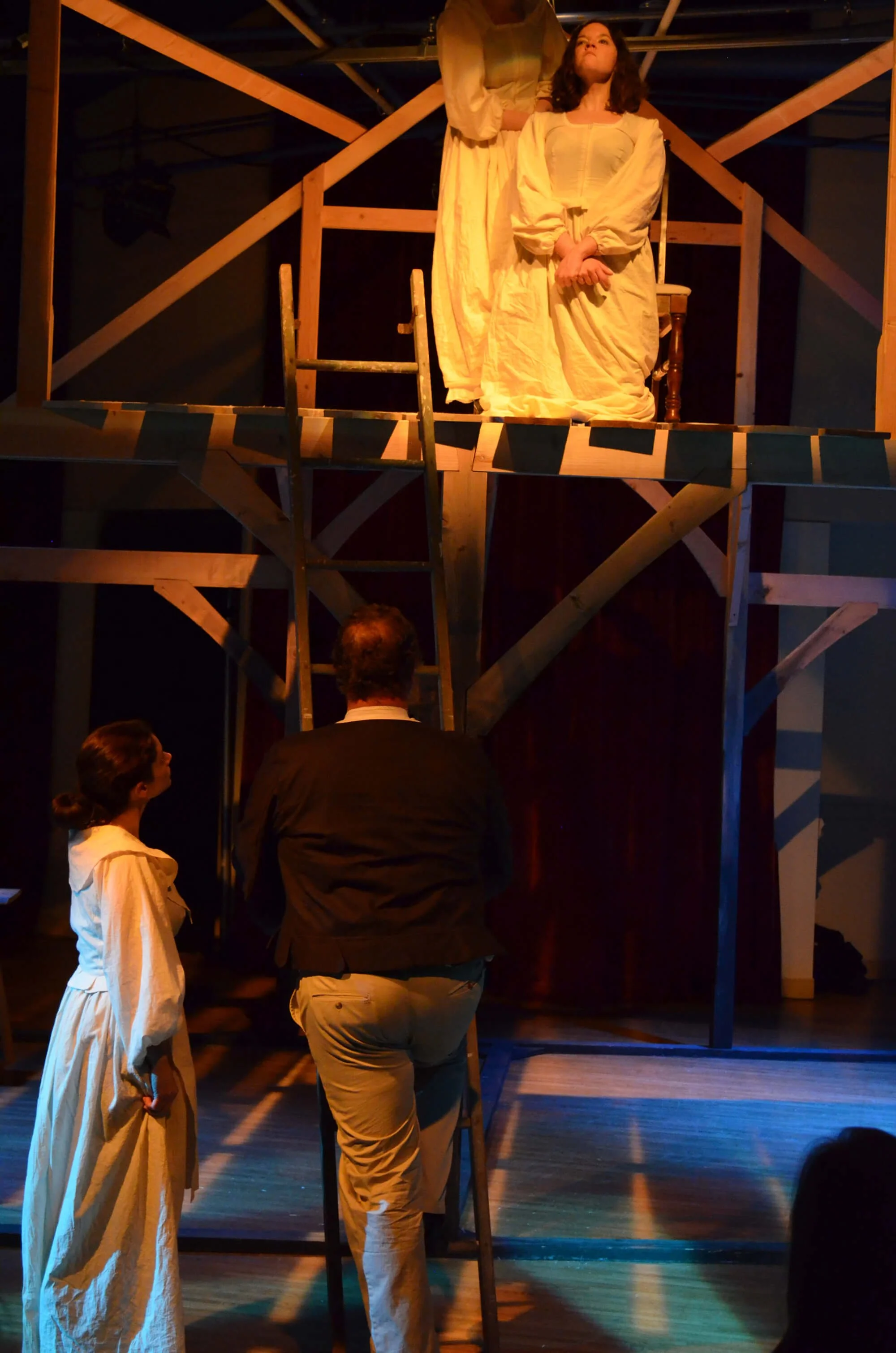 Stage scene from a theatrical play, showing a woman in a light yellow dress siting on a chair, with a man and a woman standing below, looking up at her, on a set with wooden scaffolding and stairs.