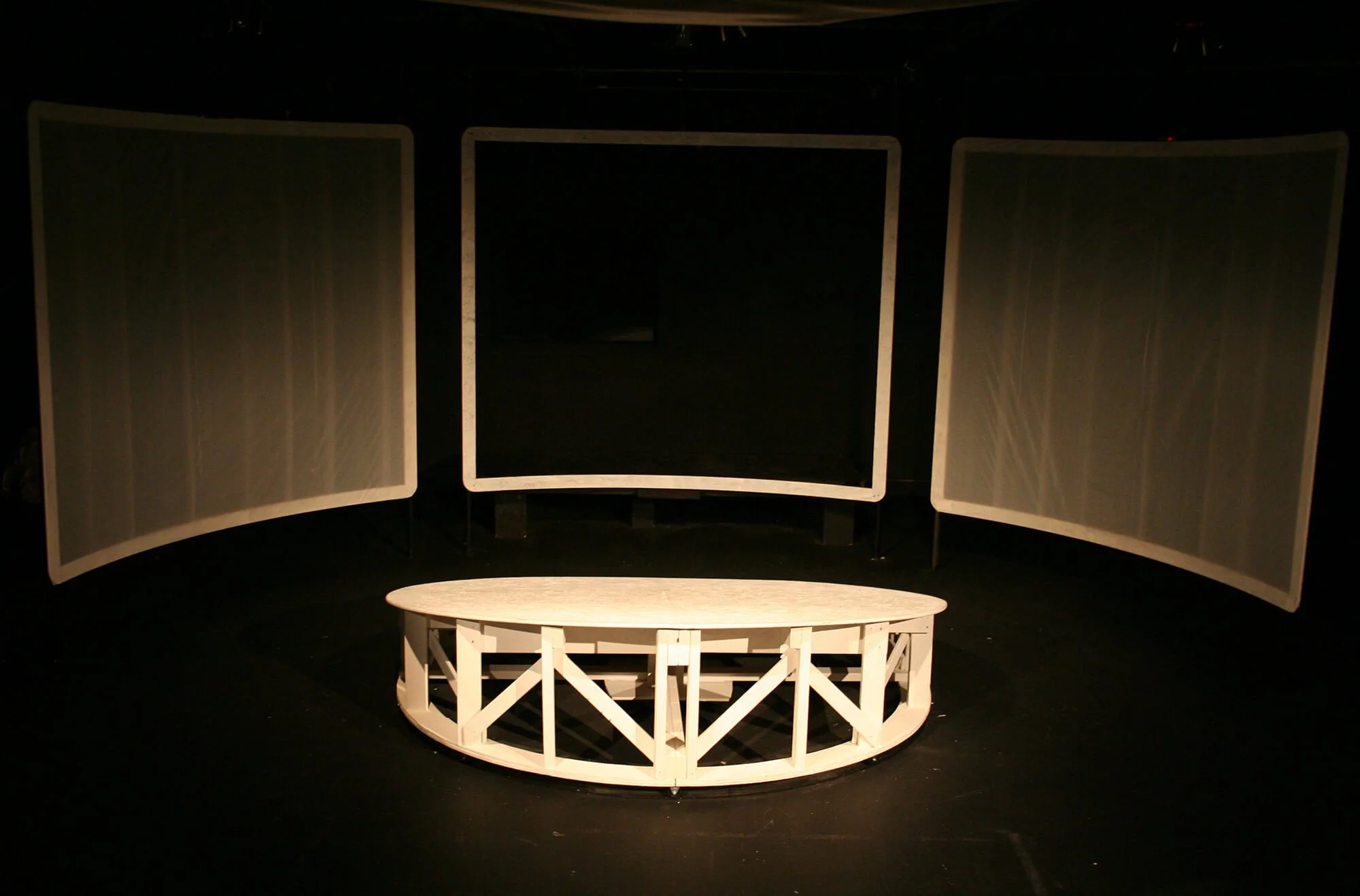 Empty stage with three vertical white screens and a white circular table in front, all set against a dark background.