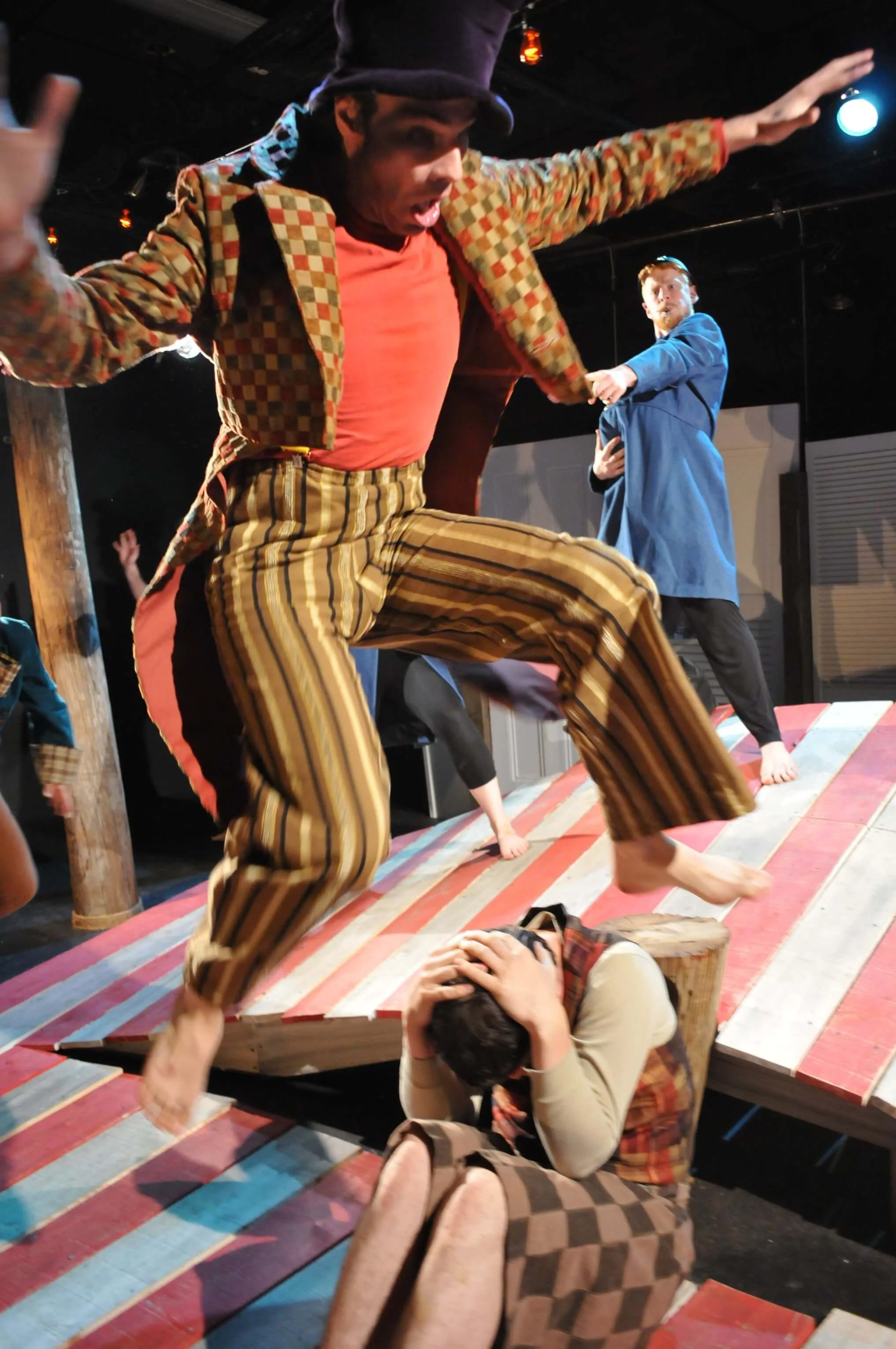 Actors performing on a stage, one in colorful striped pants and checkered jacket stomping on a person lying on the ground, who is holding their head. The stage has red, white, and gray striped wooden planks.