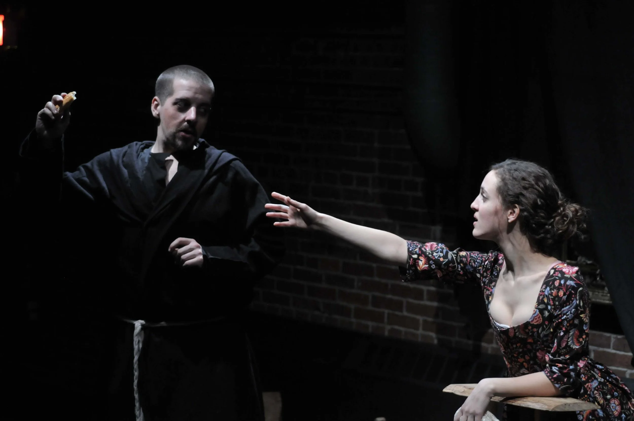 A scene from a theatrical performance showing a man in black robes holding an object in one hand, and a woman reaching out to him with her arm extended. The woman has curly hair and is wearing a floral dress. The background is dark with a brick wall.