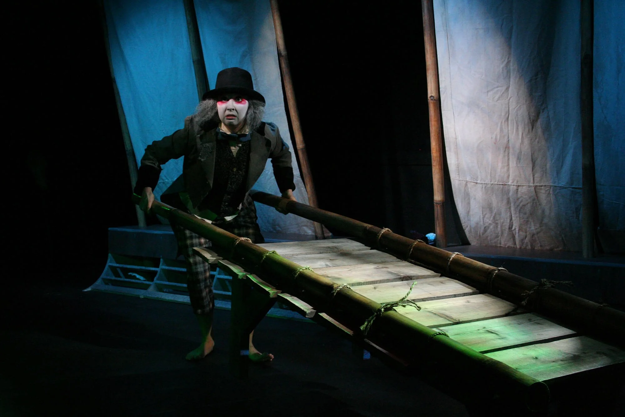 A person on a stage dressed in a clown costume with a painted face, wearing a black top hat, tuxedo, and plaid pants, holding a bamboo pole, with a makeshift wooden bridge and fabric backdrop behind them.