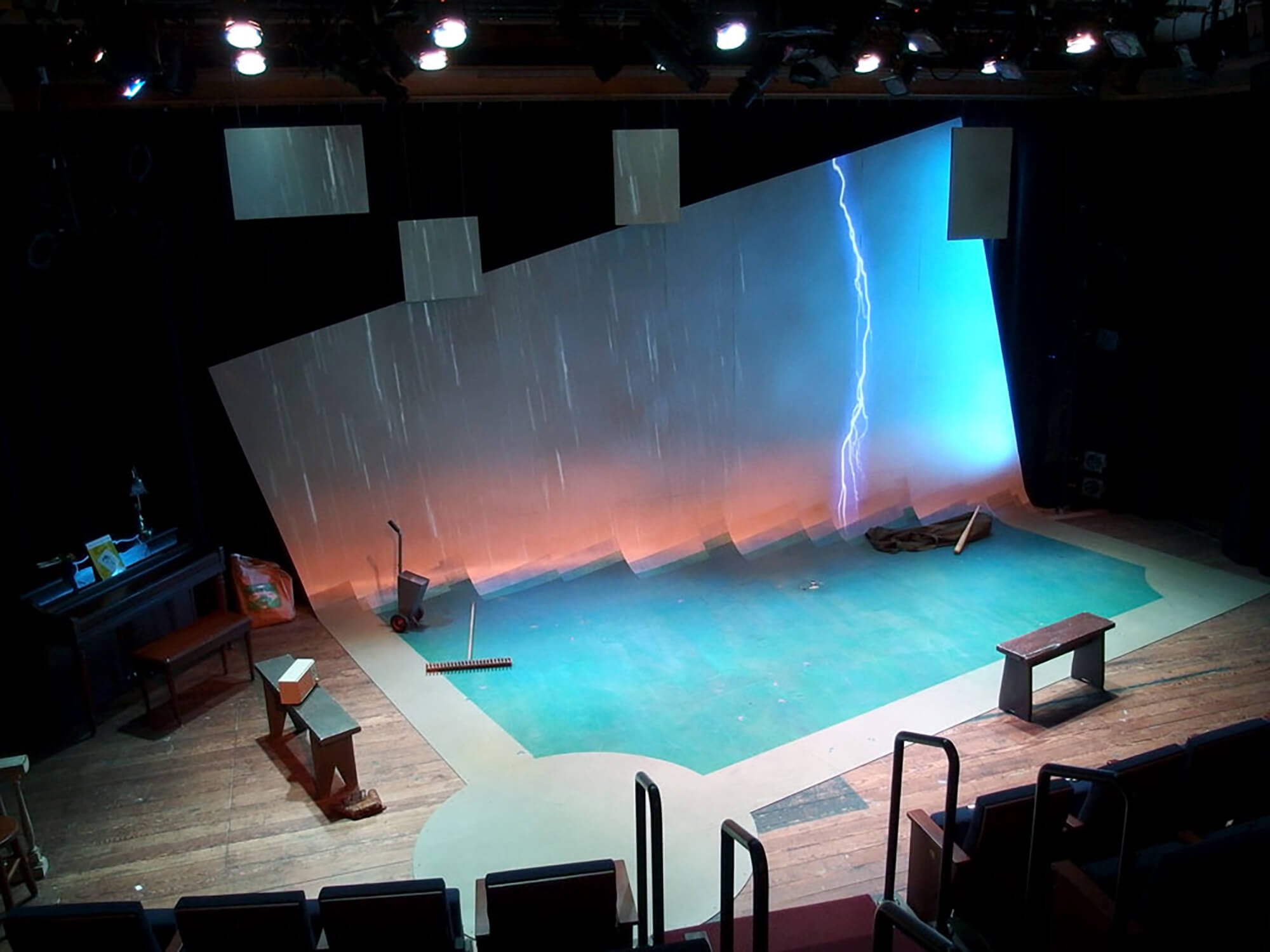 Empty stage with a backdrop showing lightning and a cloudy sky, surrounded by benches and stage equipment, in a theater or performance space.